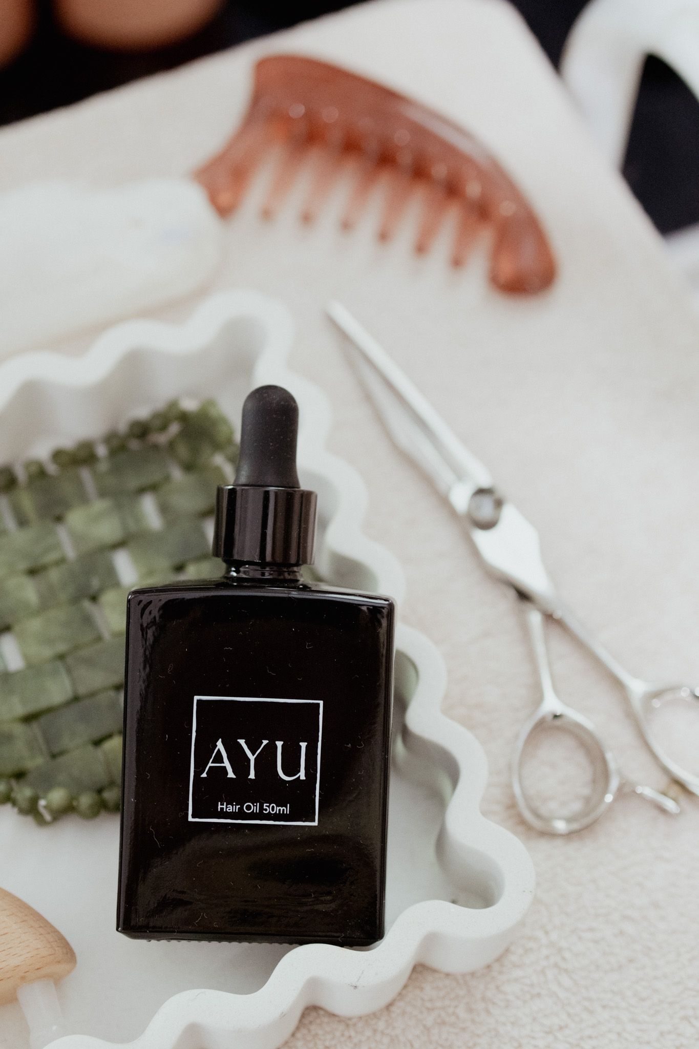 AYU hair oil 50ml in a black glass dropper bottle on a white wavy tray with hair scissors, a gua sha tool and an amber comb.