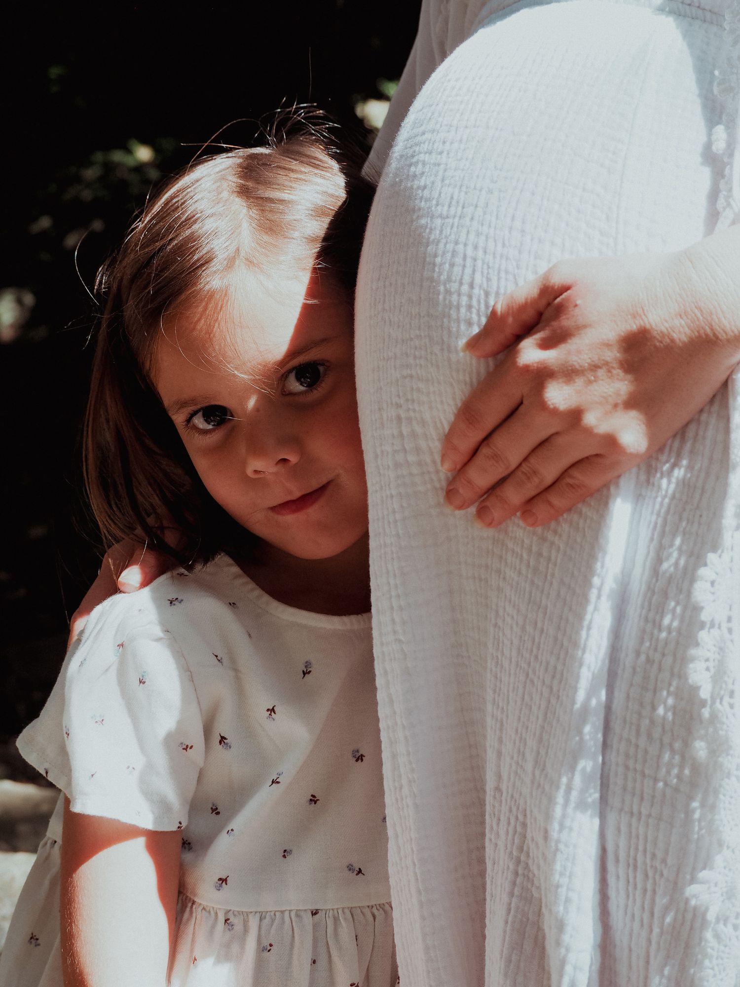 Close-up of child peeking out beside person in white textured dress in sunlit setting.