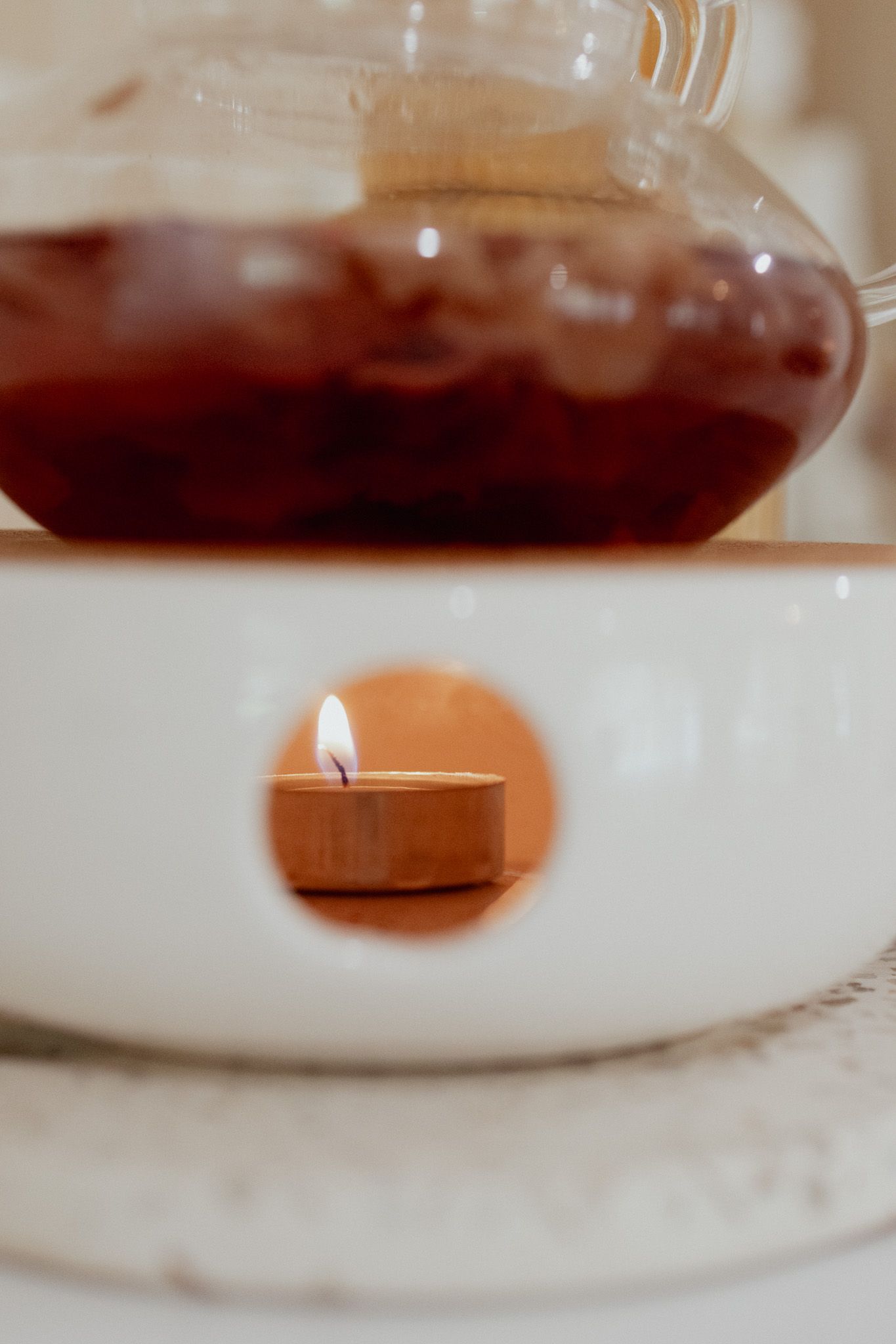A lit tealight candle glows inside a white ceramic warmer beneath a glass teapot filled with steeping red fruit tea.