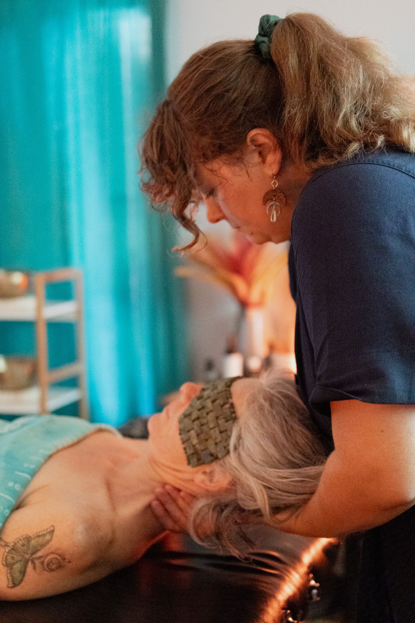 A massage therapist gently cradles a client's neck and head in a calm spa room, the client wearing a woven eye mask and a butterfly tattoo.