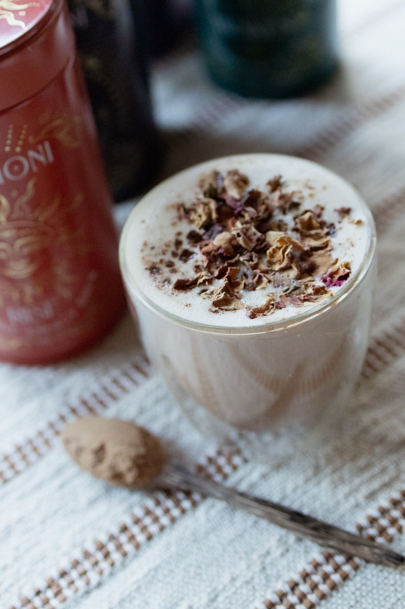 Frothy latte in a double-walled glass topped with dried rose petals, beside a spoon of powder and a red tin on a woven linen cloth.