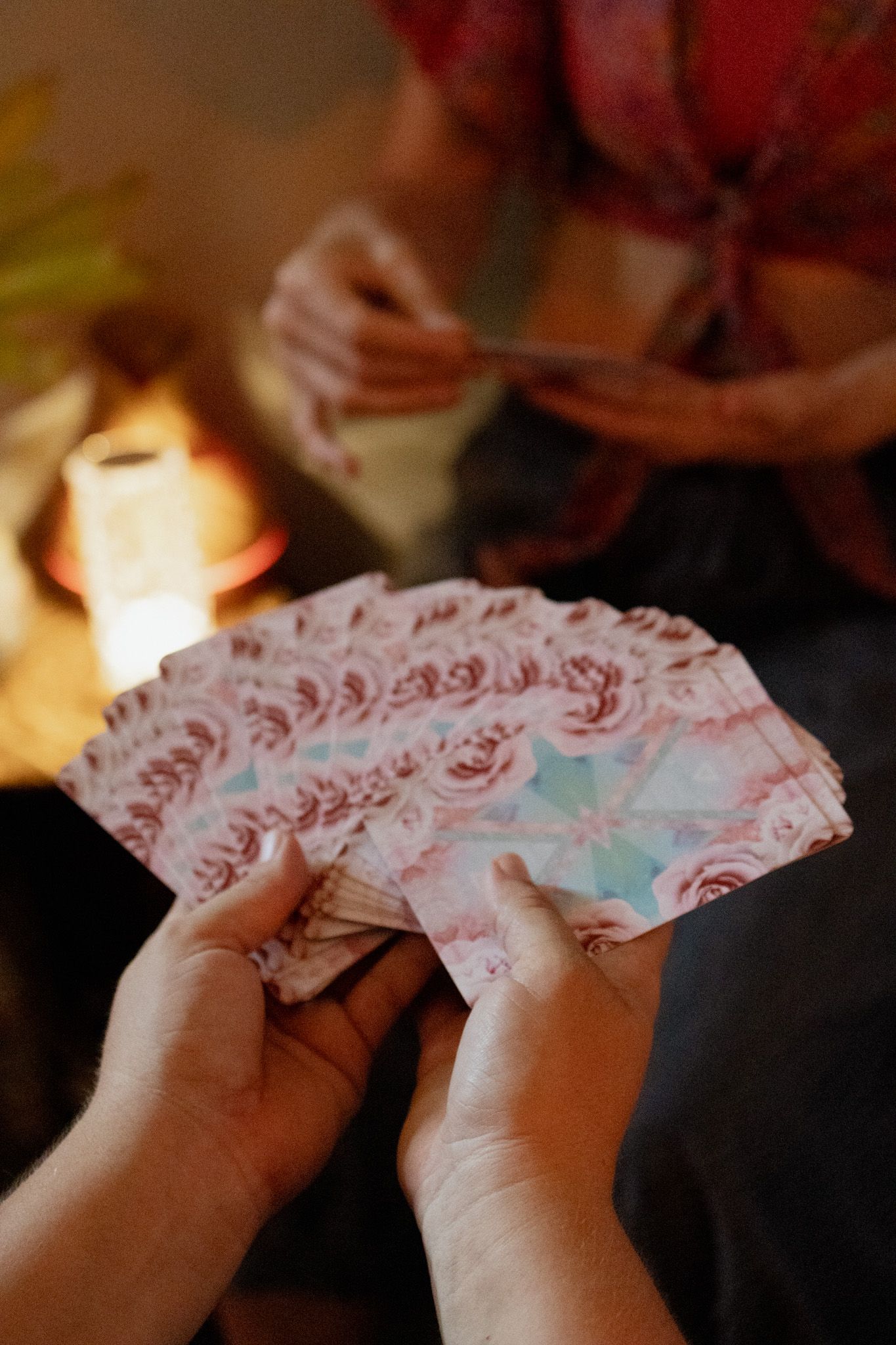 Hands fanning out a deck of pink floral oracle cards during a tarot reading, with candlelight glowing softly in the background.