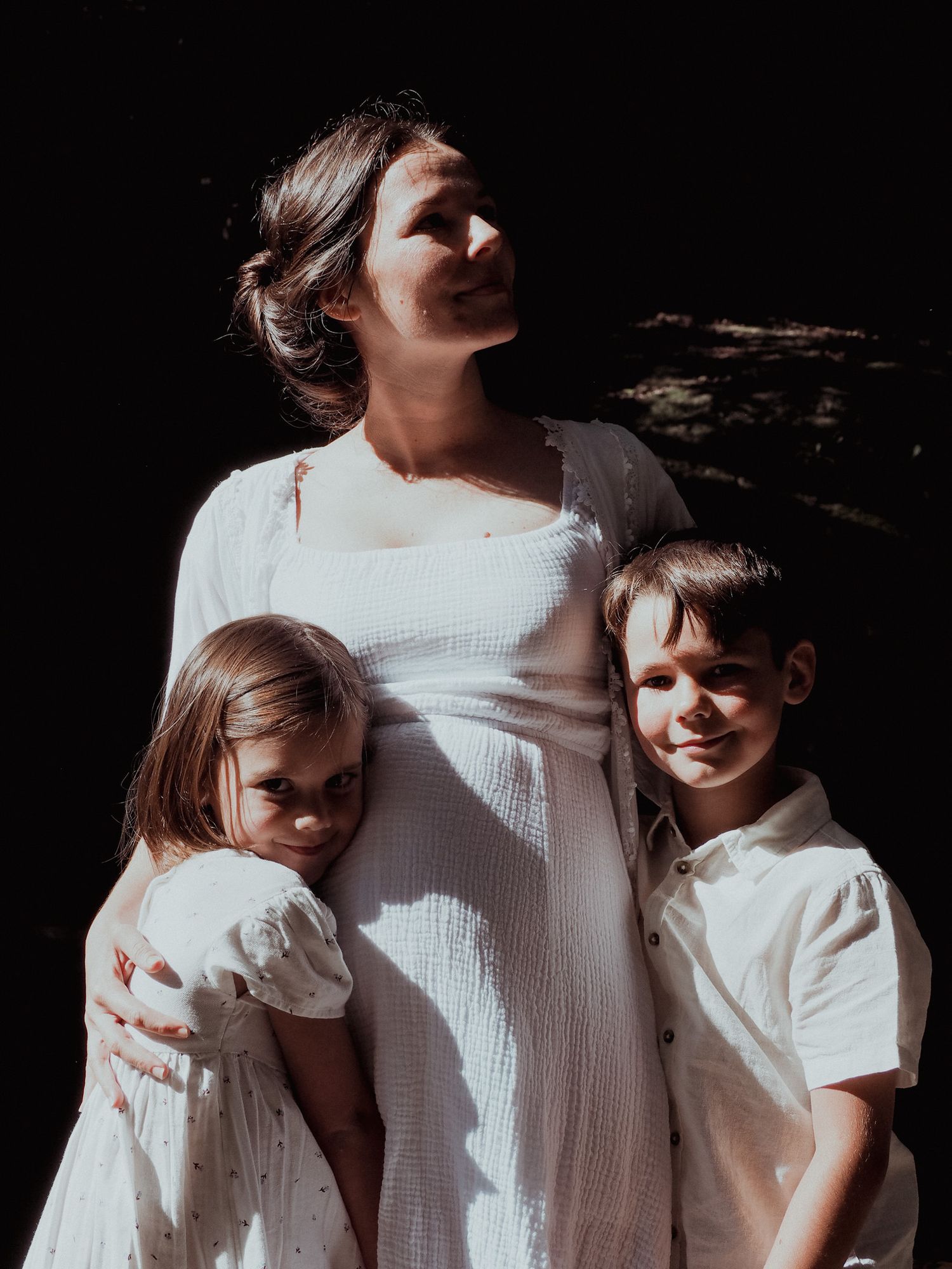 Family portrait showing tender moment between parent and children in white outfits against dark background.
