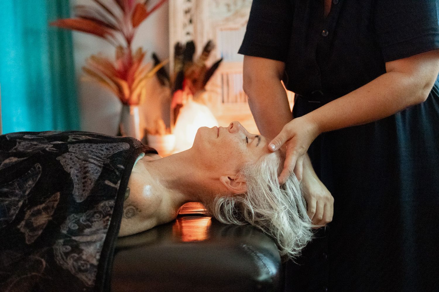 A gray-haired woman lies relaxed on a massage table while a therapist gently massages her neck and head in a warm, candlelit spa room.