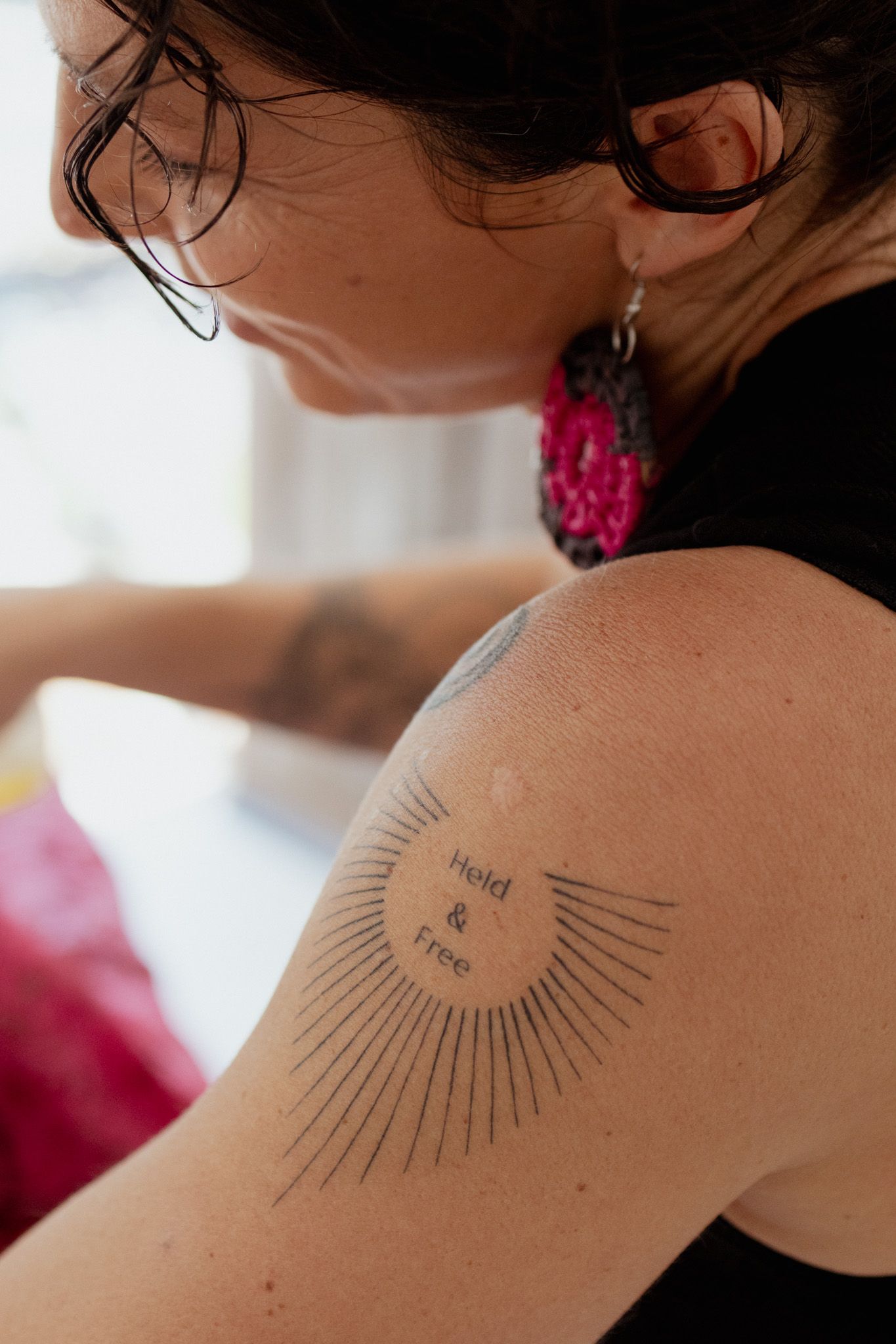 Woman with glasses and pink crocheted earrings shows a sunburst tattoo reading 