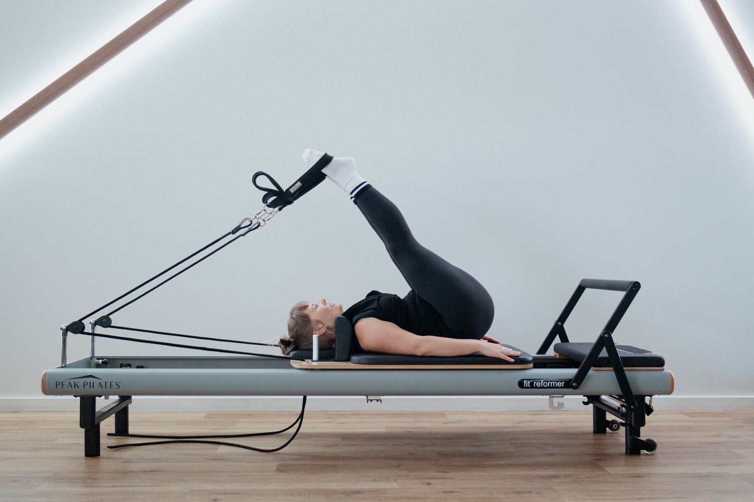 Woman lying on a Pilates reformer in a bright studio with one leg raised and strapped to the springs during a core and hamstring exercise.