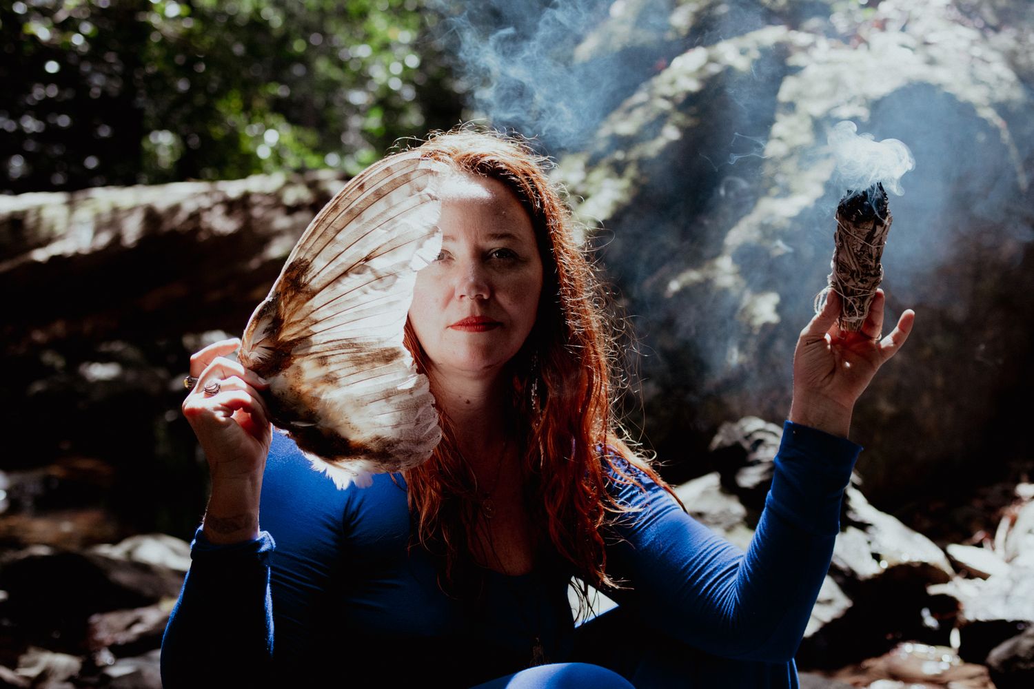 Person with long hair holding burning sage in blue sweater against rocky background.