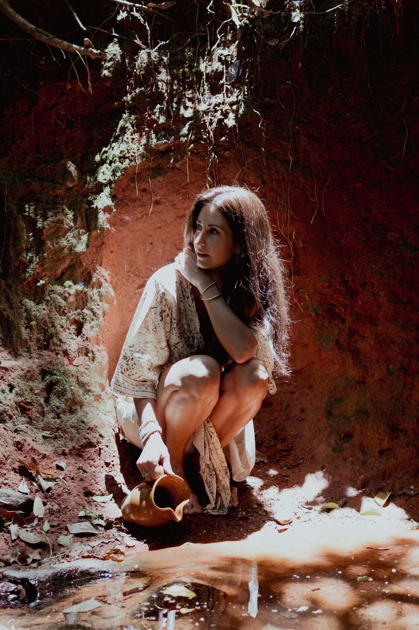 Person in white dress crouches dramatically against a red clay background in nature.