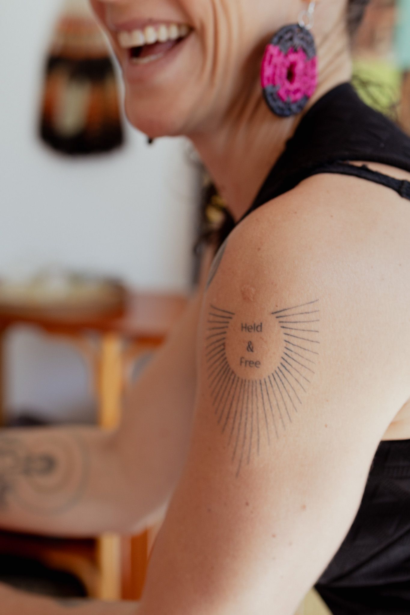Smiling woman with pink beaded earrings shows a sunburst tattoo reading 