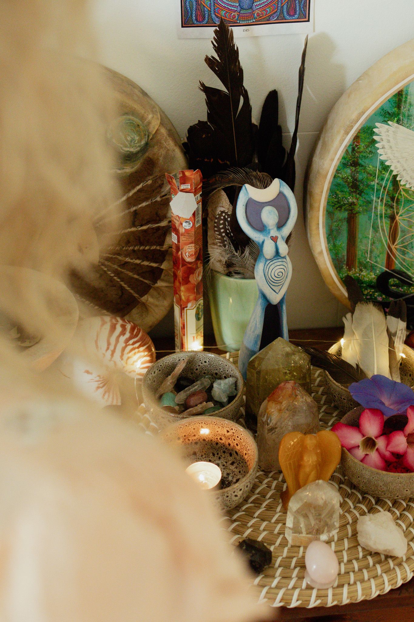 Spiritual altar with a goddess figurine, black feathers, crystals, seashells, candles, and pink plumeria flowers on a woven tray.