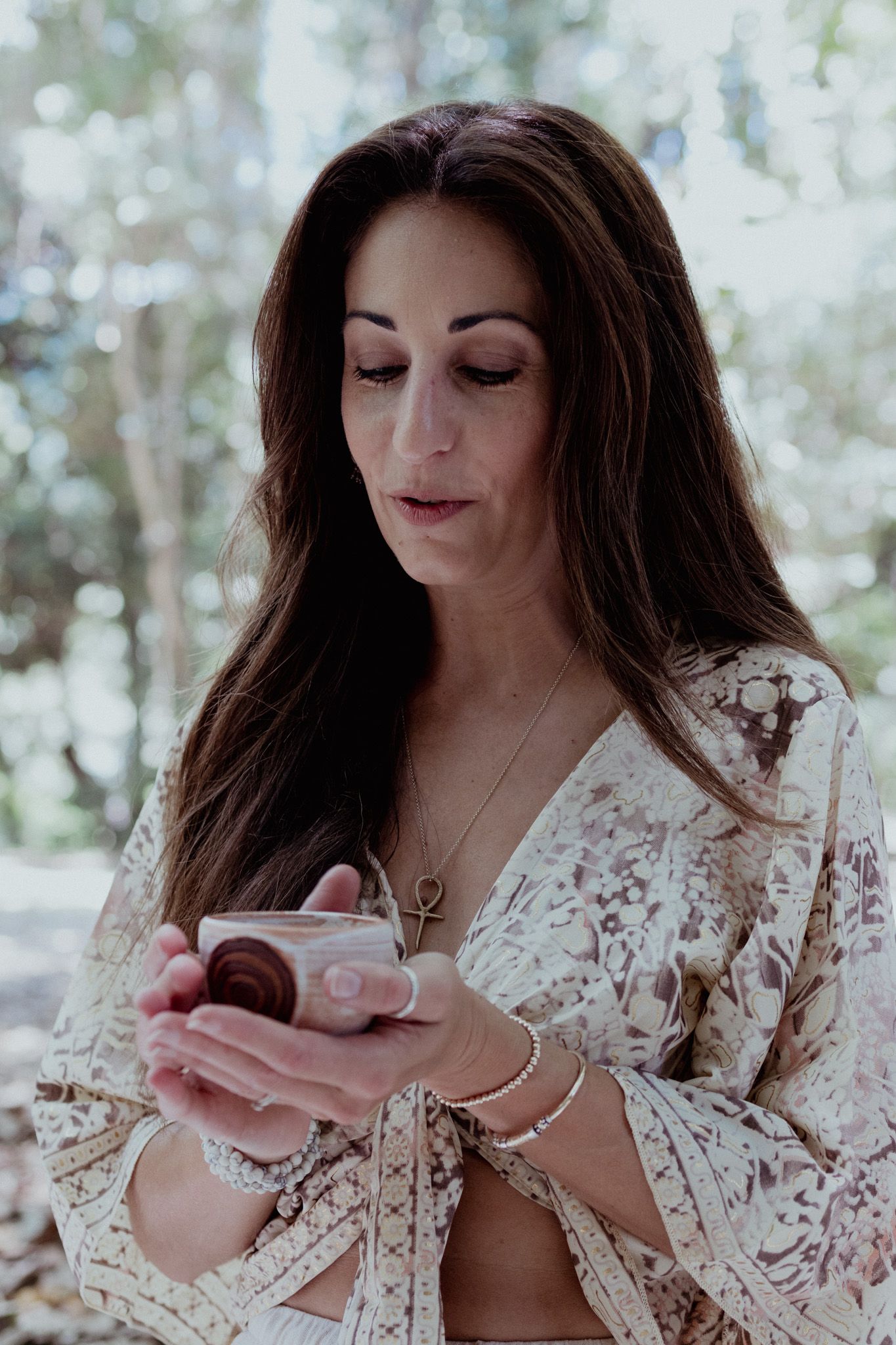 Person in lace robe holding teacup in outdoor forest setting with soft bokeh lighting.