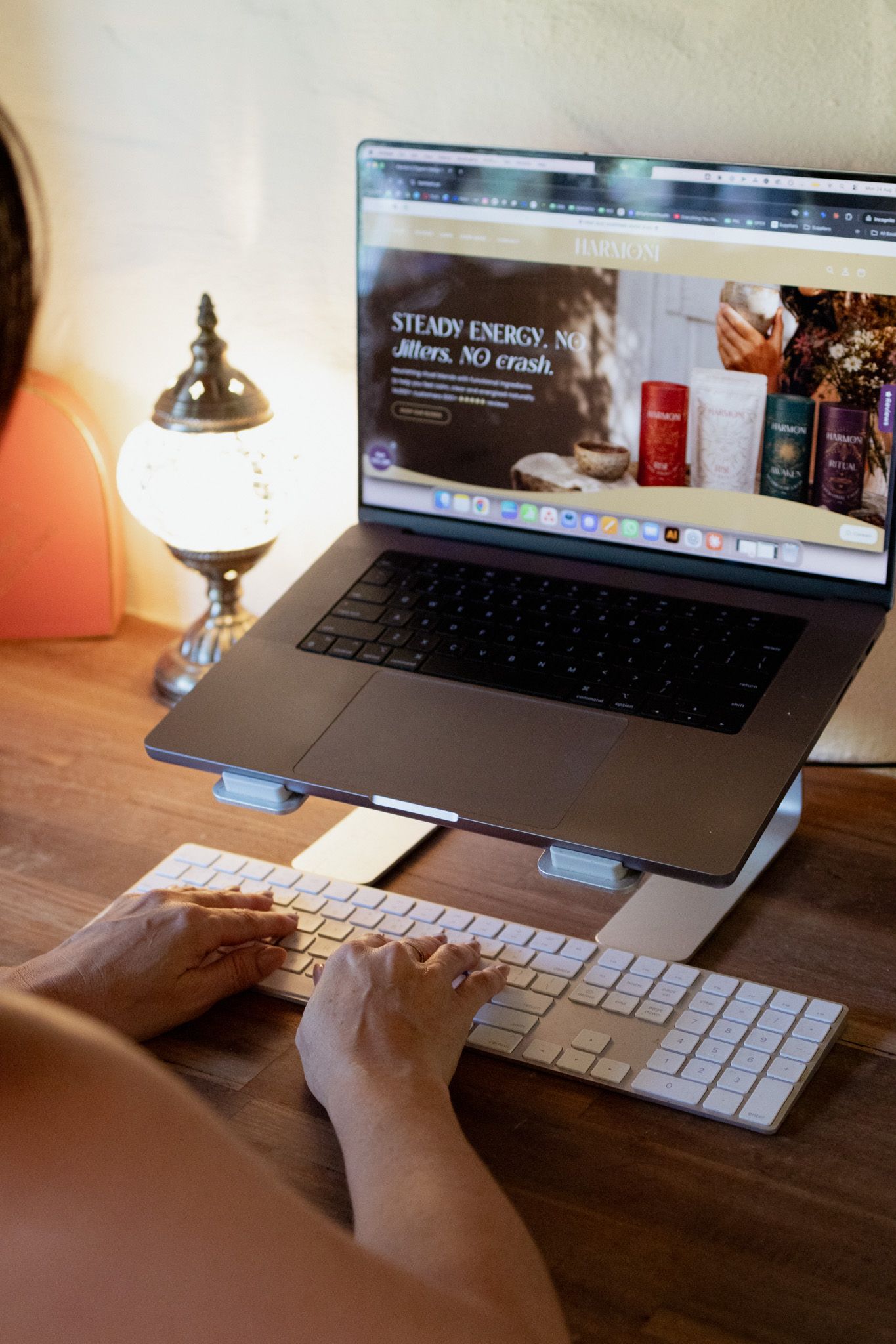 Person typing on a wireless keyboard beside a laptop on a stand showing a wellness website, with a glowing mosaic lamp on a wooden desk.