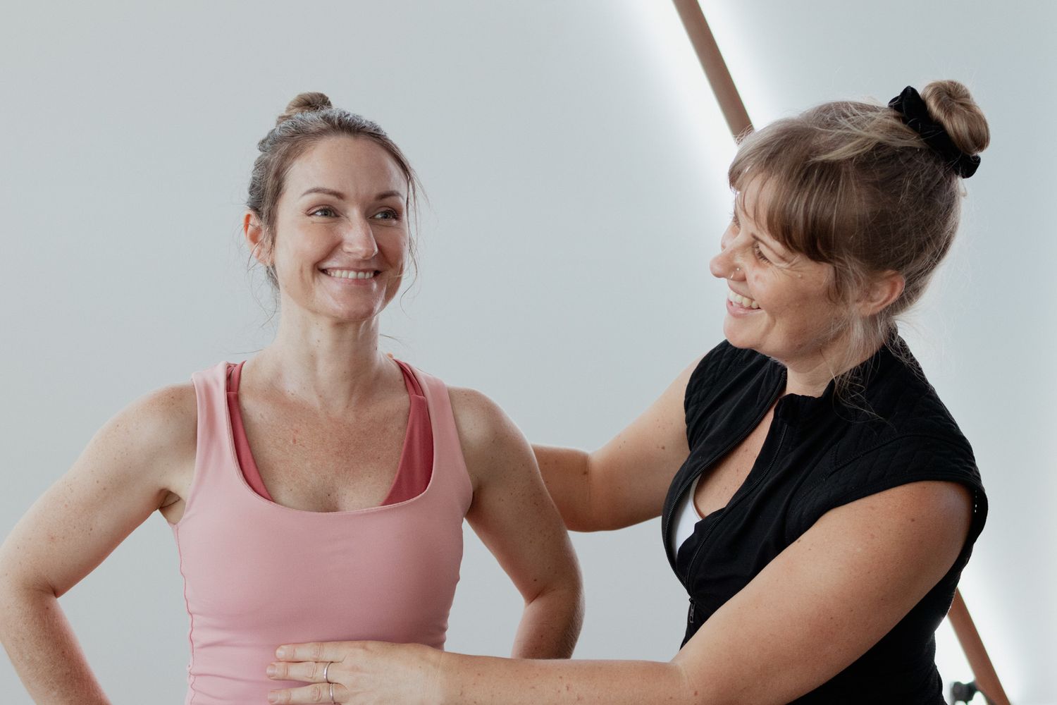 A smiling woman in a pink tank top shares a warm moment with her Pilates instructor in a bright, modern studio.