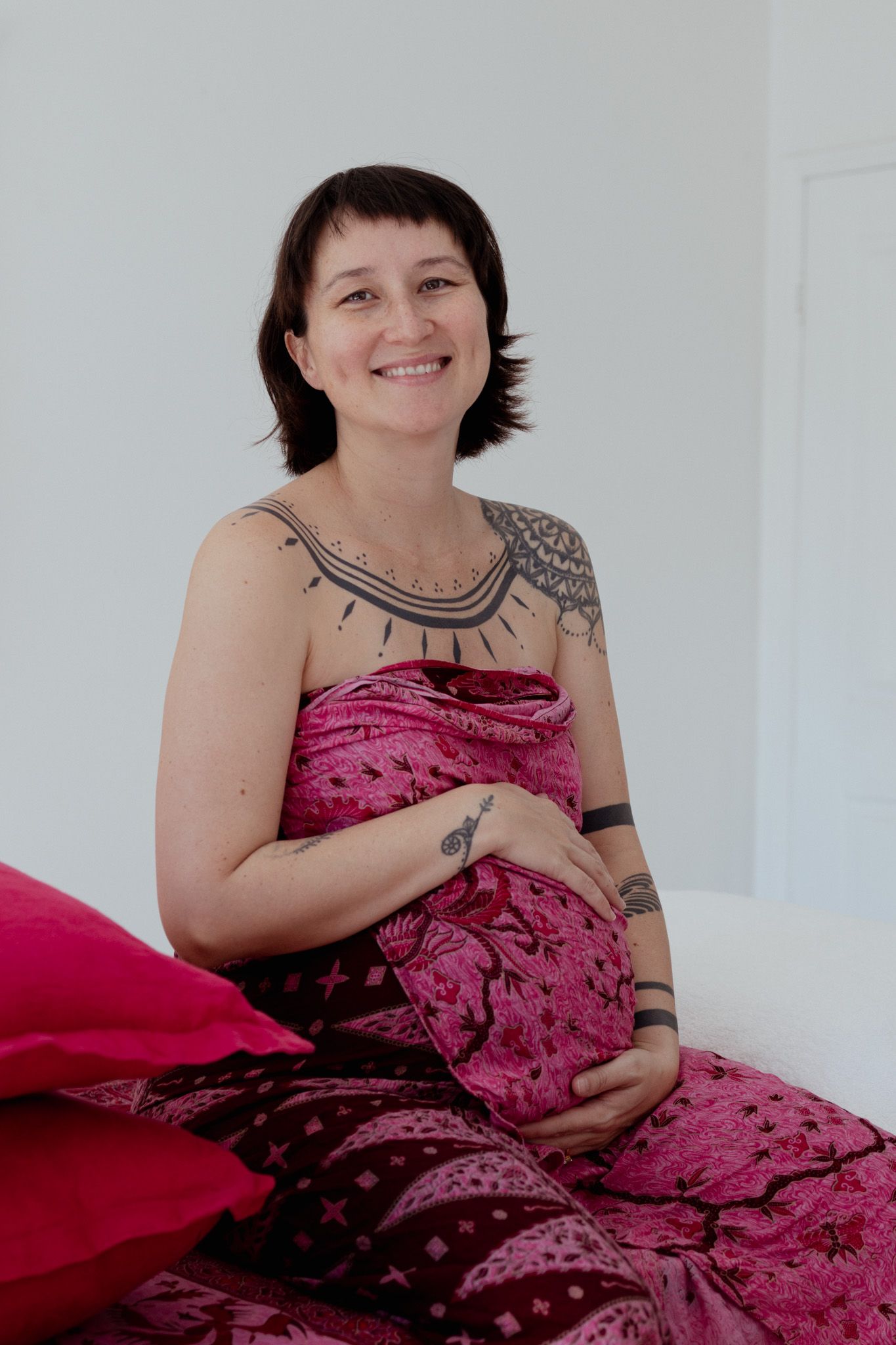 Smiling pregnant woman with short dark hair and chest tattoos sits holding her belly, wrapped in a pink patterned cloth beside red cushions.