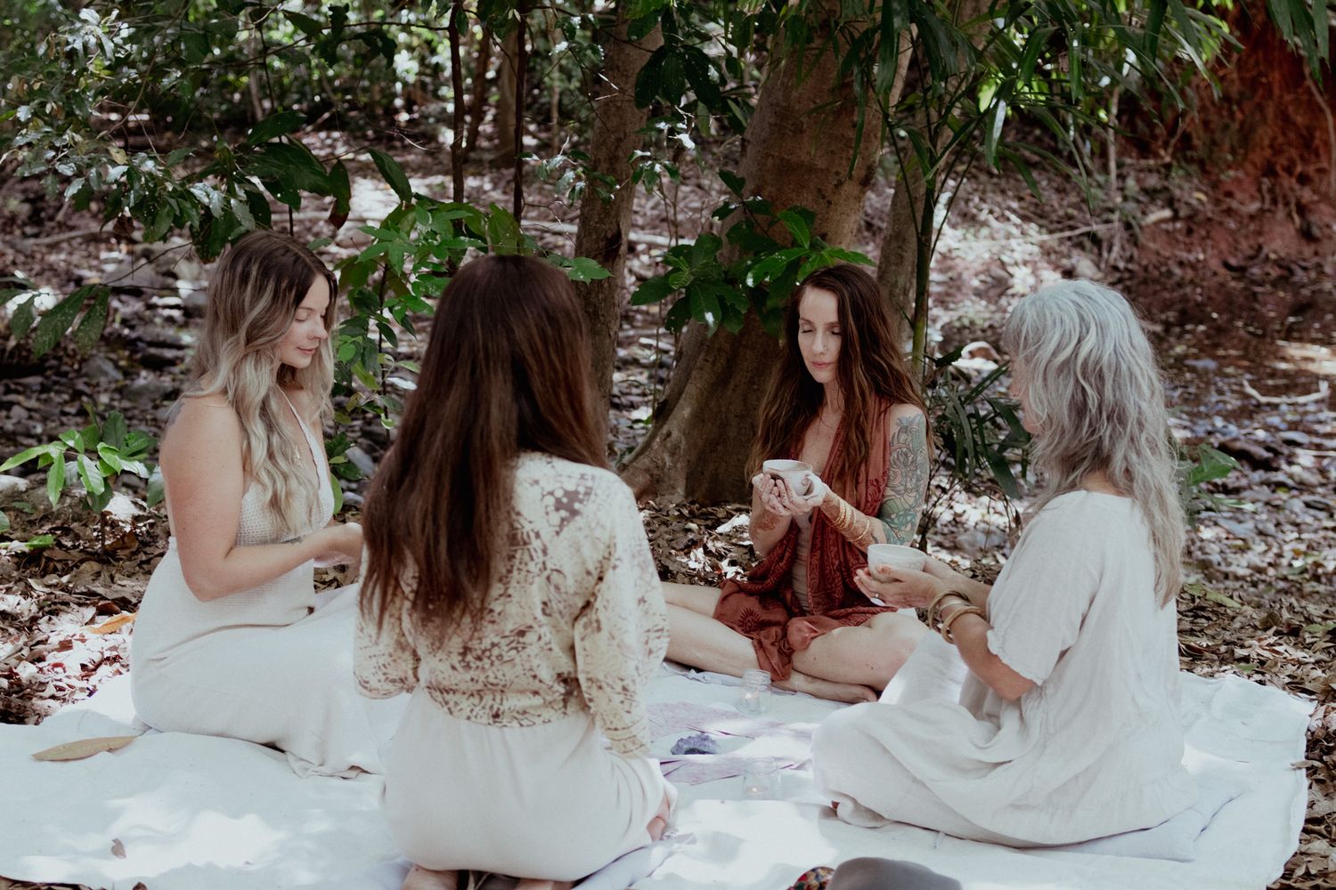 A group in white dresses sits together in a wooded outdoor setting creating an ethereal scene.