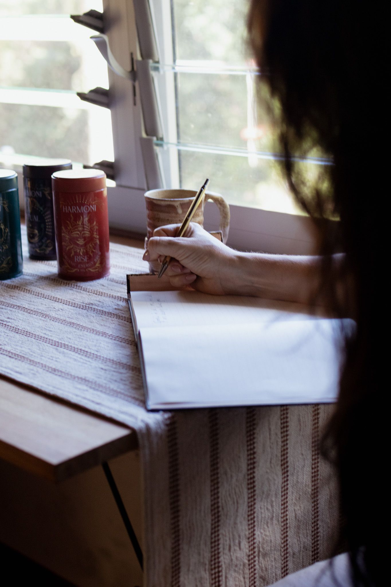 Woman with long dark hair writes in an open notebook at a wooden desk beside a sunlit window, with a ceramic mug and tea tins nearby.