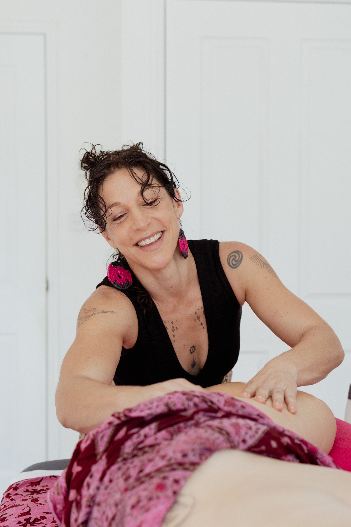 Smiling massage therapist in a black top with pink earrings gives a belly massage to a client covered in pink patterned cloth.