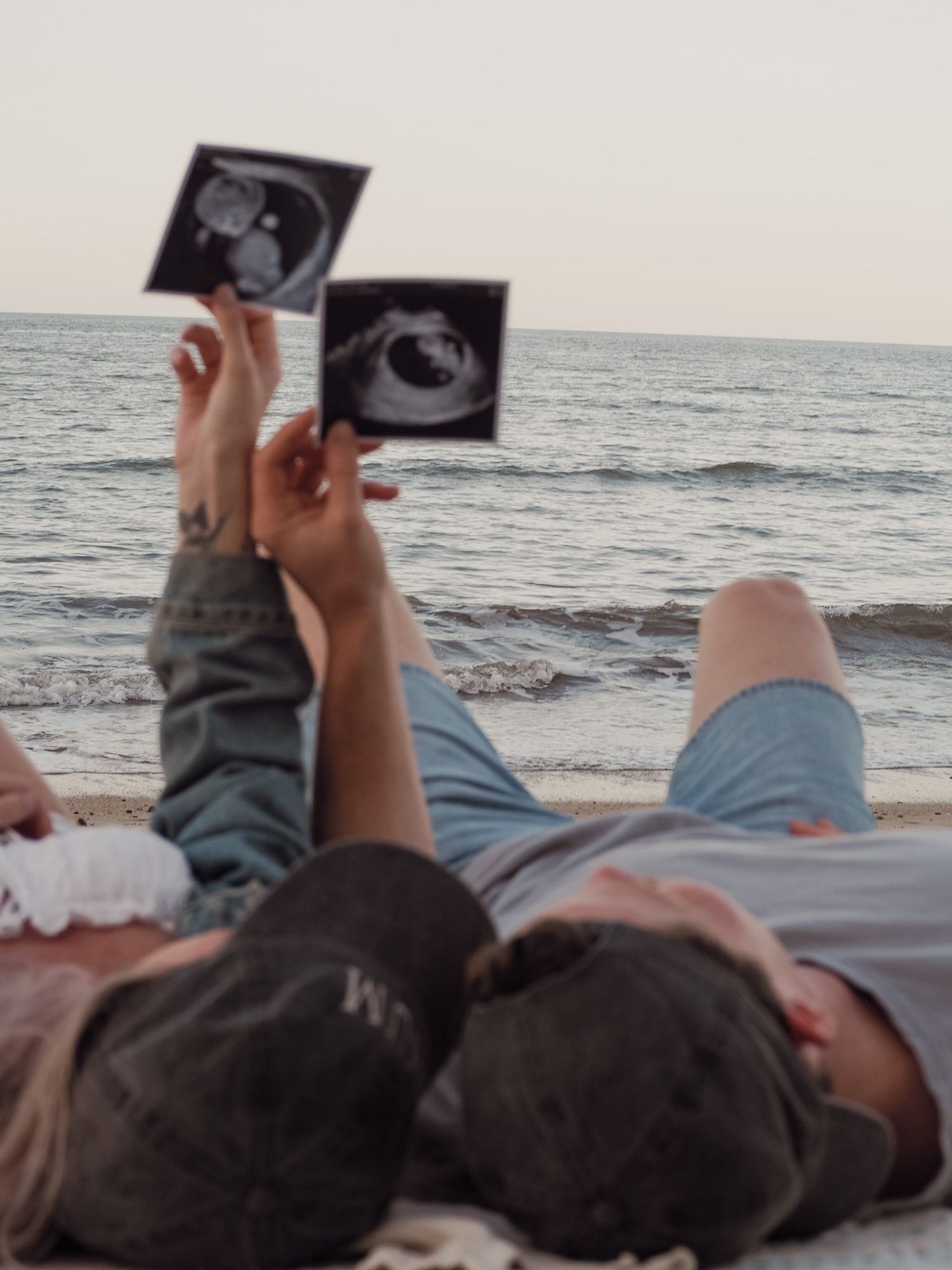 Couple lying on beach holding ultrasound photos against ocean horizon at sunset.