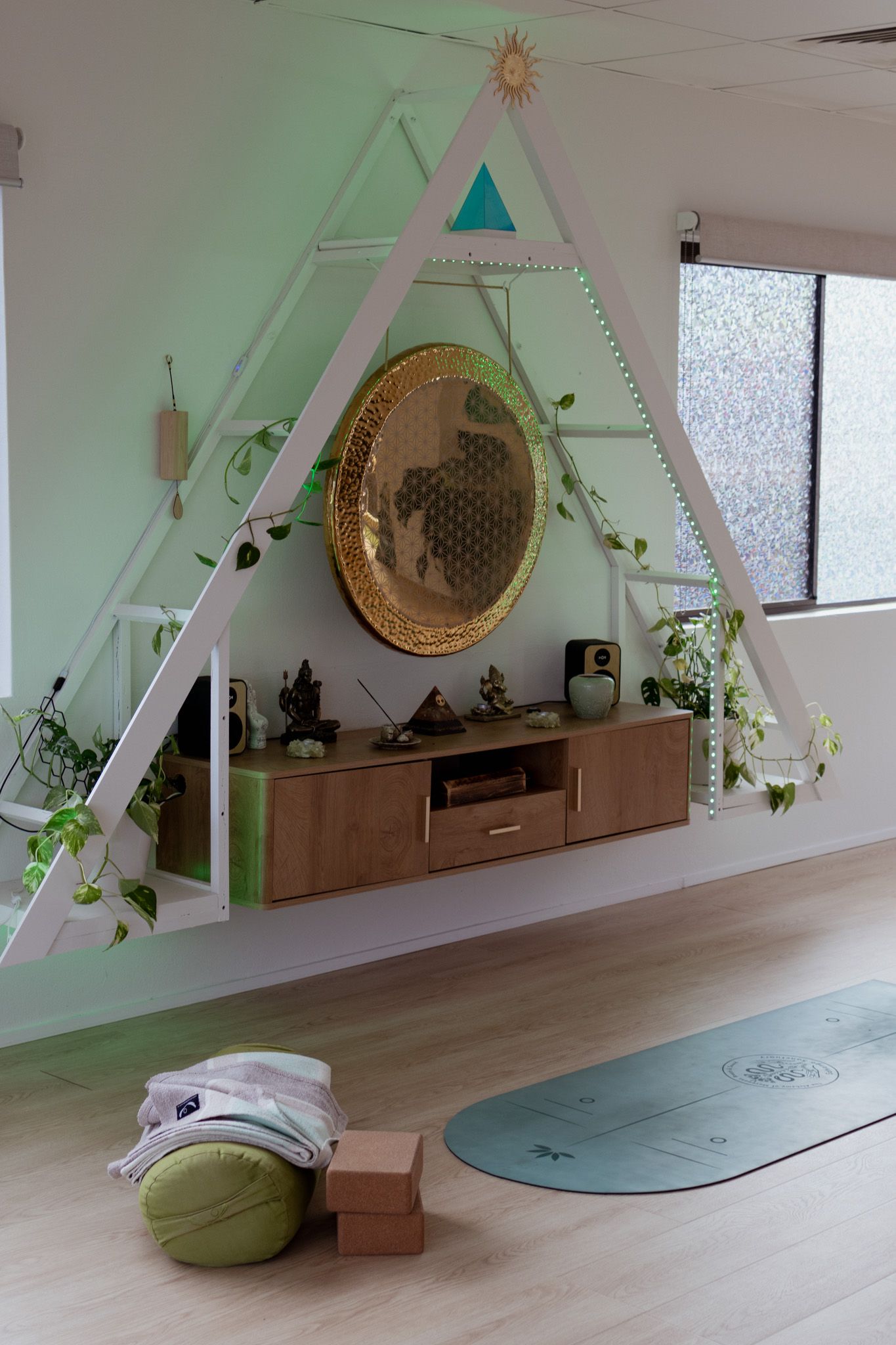 Yoga studio corner with a triangular wall shelf, gold gong, wooden floating cabinet, plants, green yoga mat, bolster and blocks.