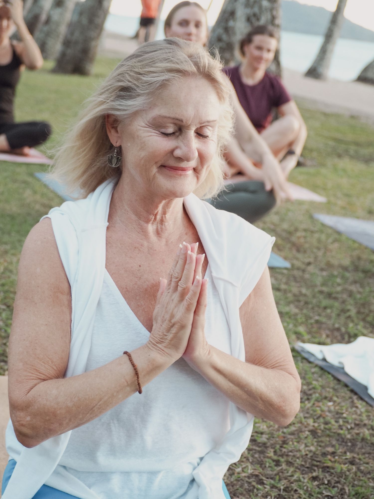 Woman in white sleeveless top practices meditation outdoors with others visible in background.