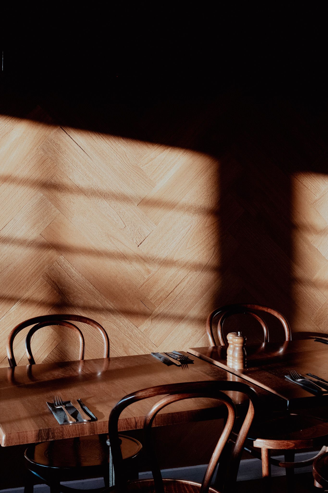 Dramatic shadows from window blinds cast across wooden dining chairs in warm sunlight.