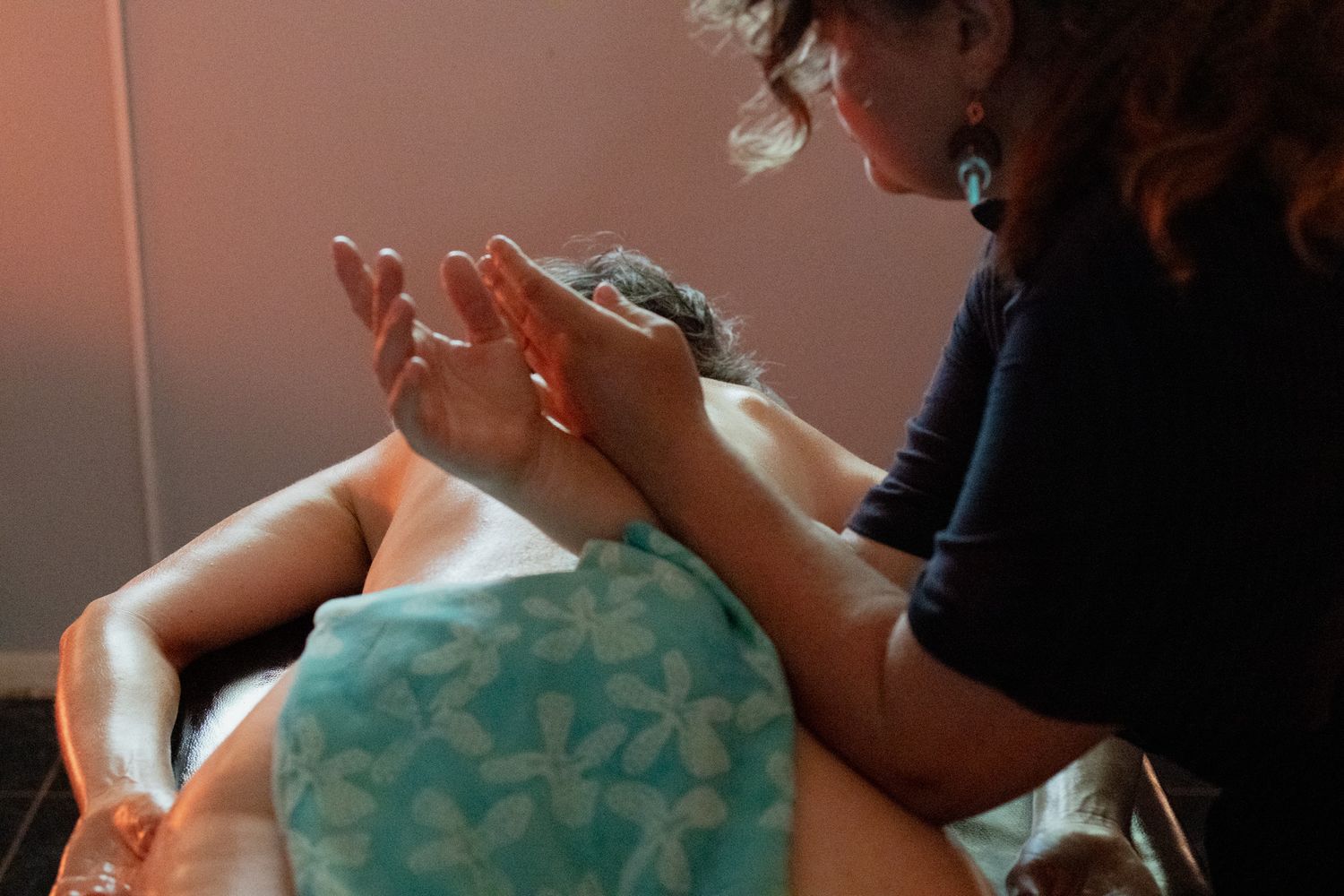 Massage therapist with curly hair performs a relaxing back massage on a client covered with a teal floral towel in a warmly lit spa room.