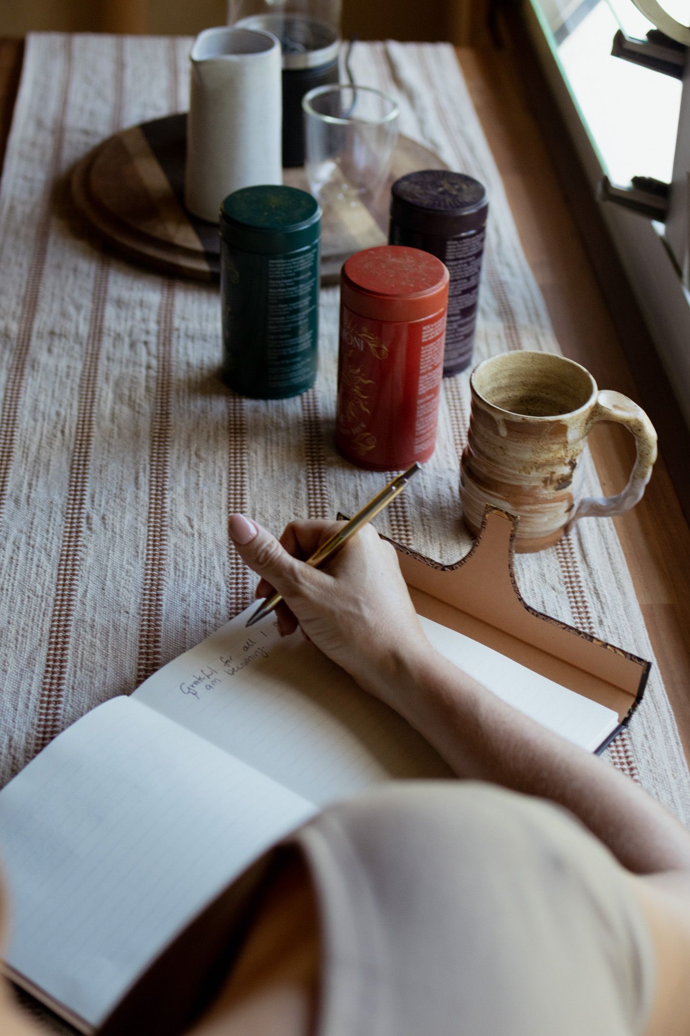 Person writing with a gold pen in an open leather journal beside a handmade ceramic mug and colorful tea tins on a woven linen table runner.