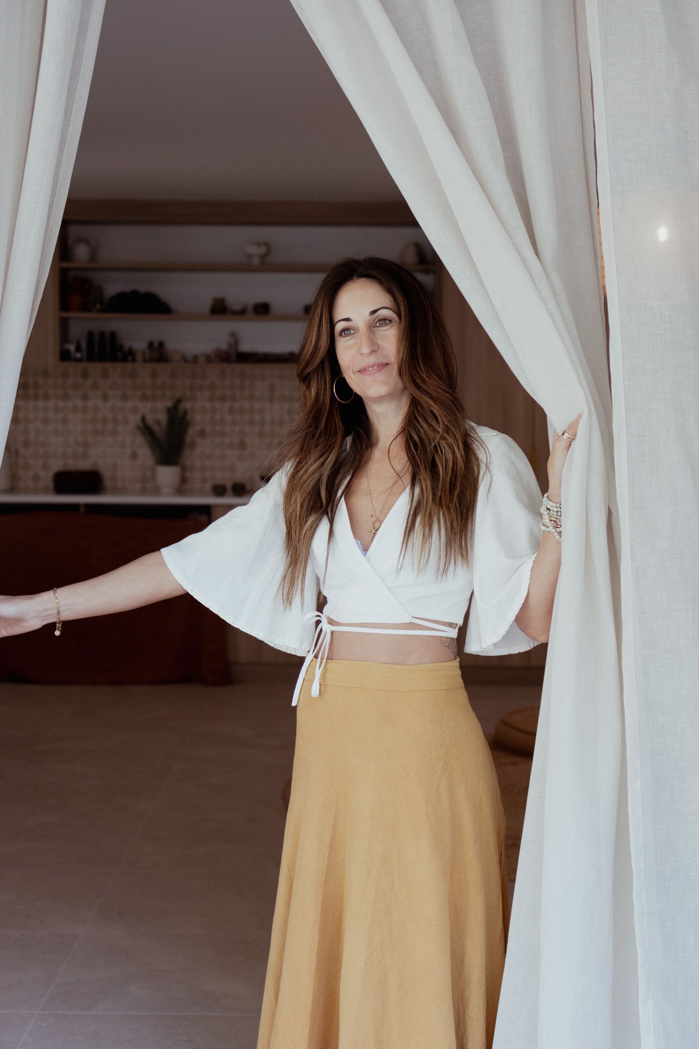 Woman with long brown hair in a white wrap top and tan skirt parts sheer white curtains in a bright, earthy-toned room.