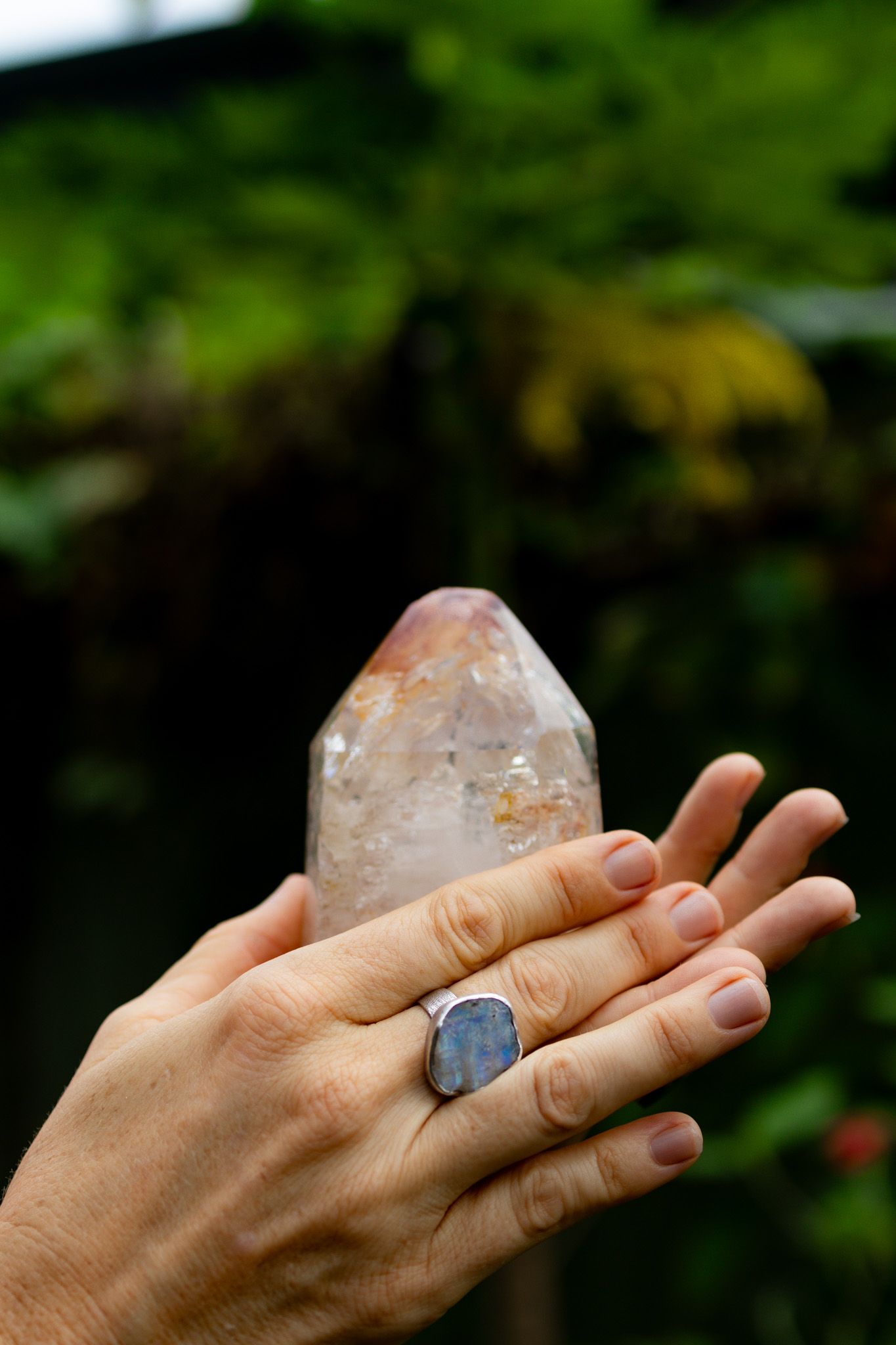 Two hands hold a large clear quartz crystal point with warm inclusions, one wearing a silver ring with a blue stone, against green foliage.