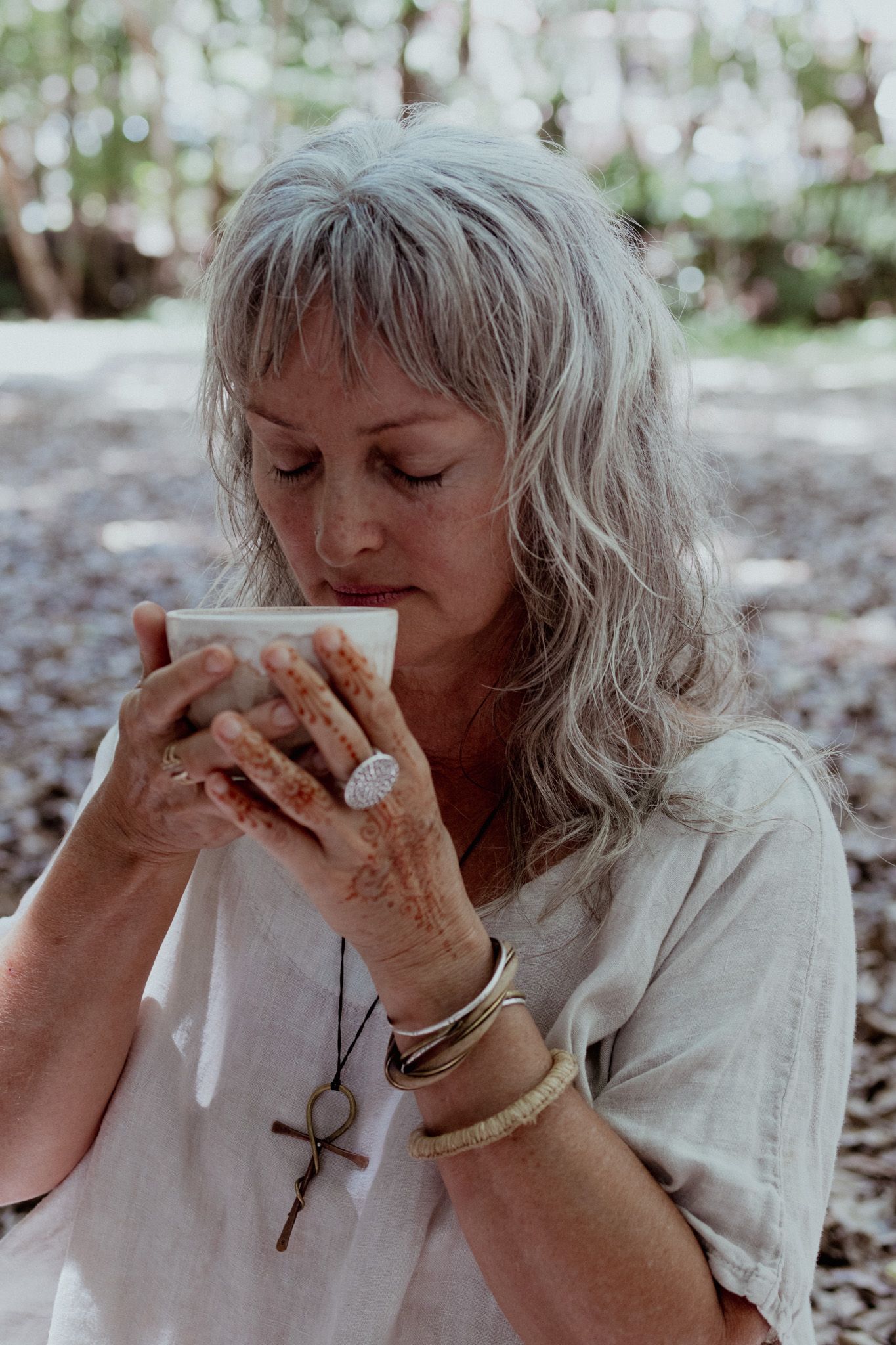Person in white clothing drinking from small cup in outdoor nature setting.