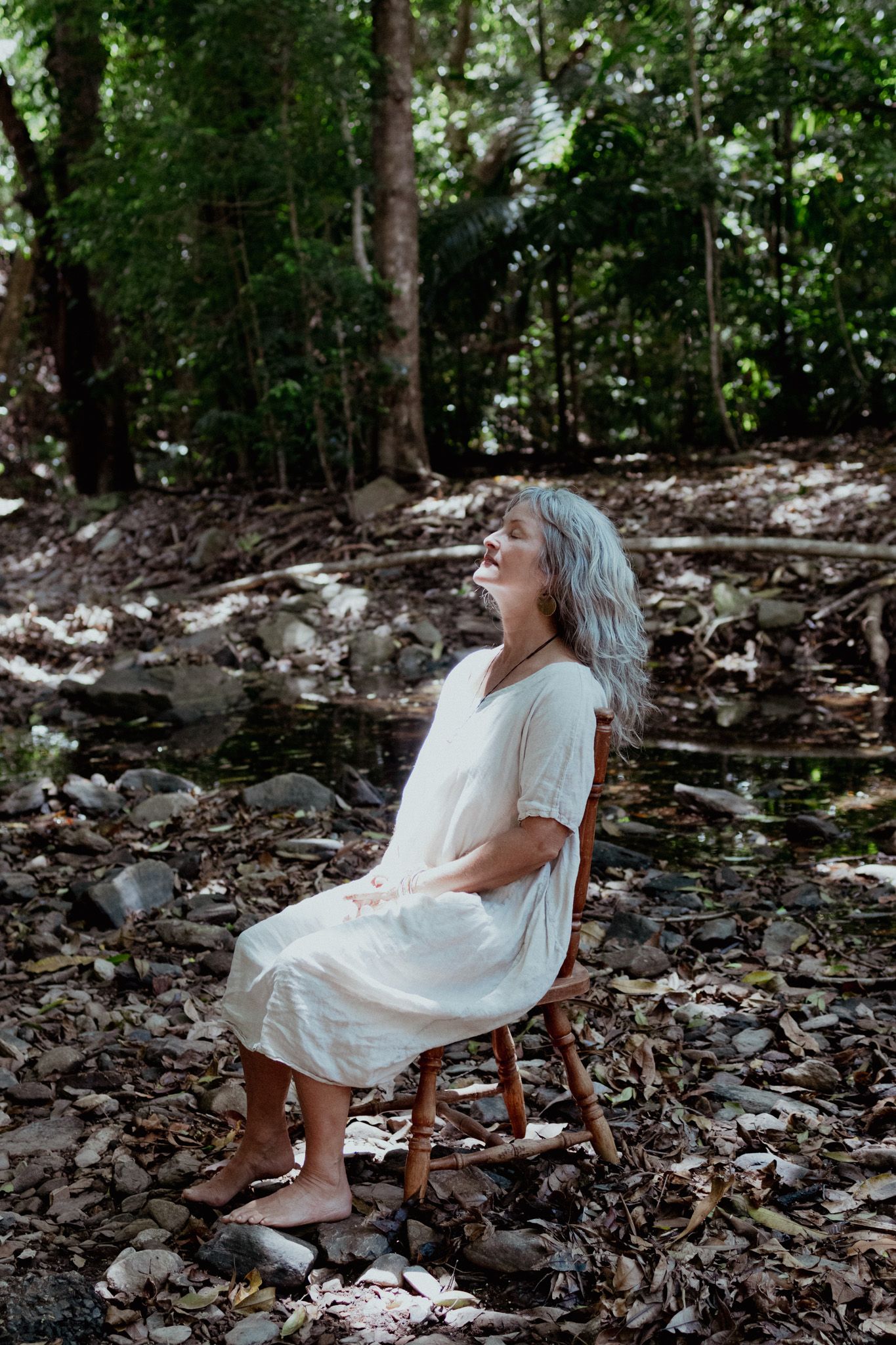 Person in white dress sitting peacefully on wooden chair in forest setting.