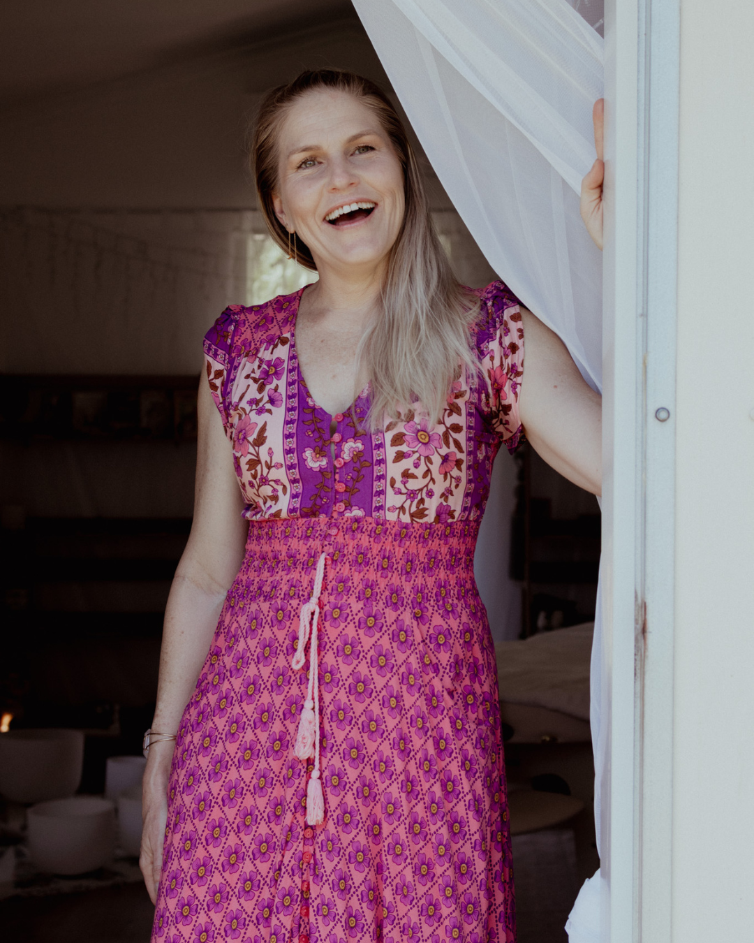 A person wearing a vibrant pink and purple floral print dress poses and smiles against a white wall.