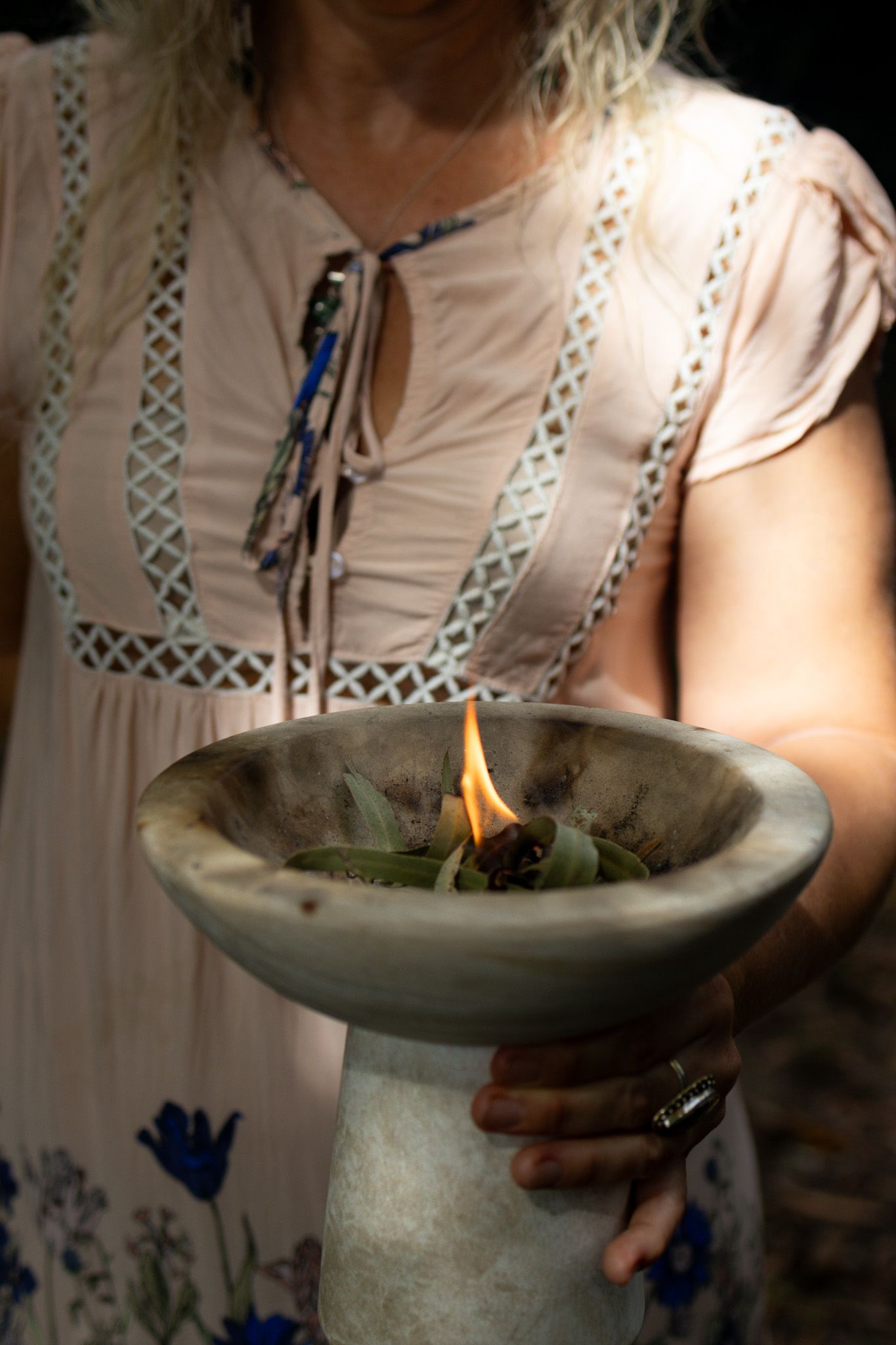 Woman in a blush pink lace-trim dress holds a stone bowl of burning sage and herbs during a smudging ritual.