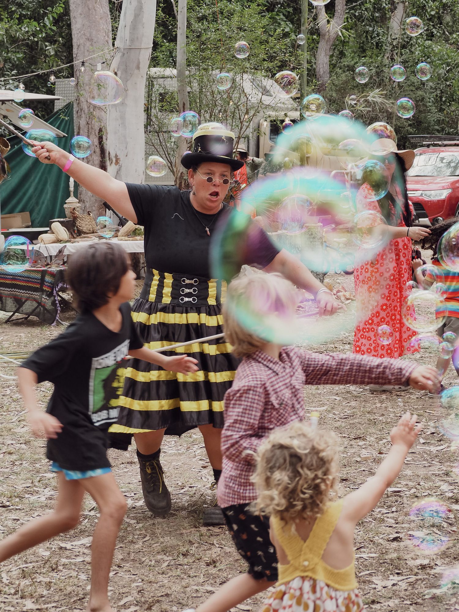 Children playing with giant soap bubbles while entertainer in striped dress performs at outdoor event.
