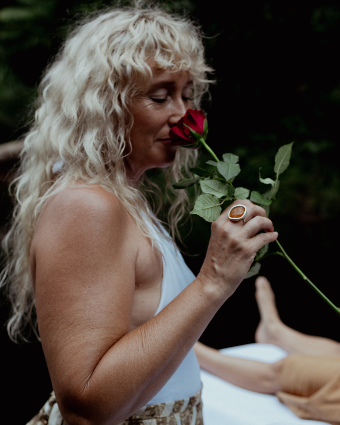 Woman with long blonde wavy hair in white top smells red rose against dark background in romantic portrait.