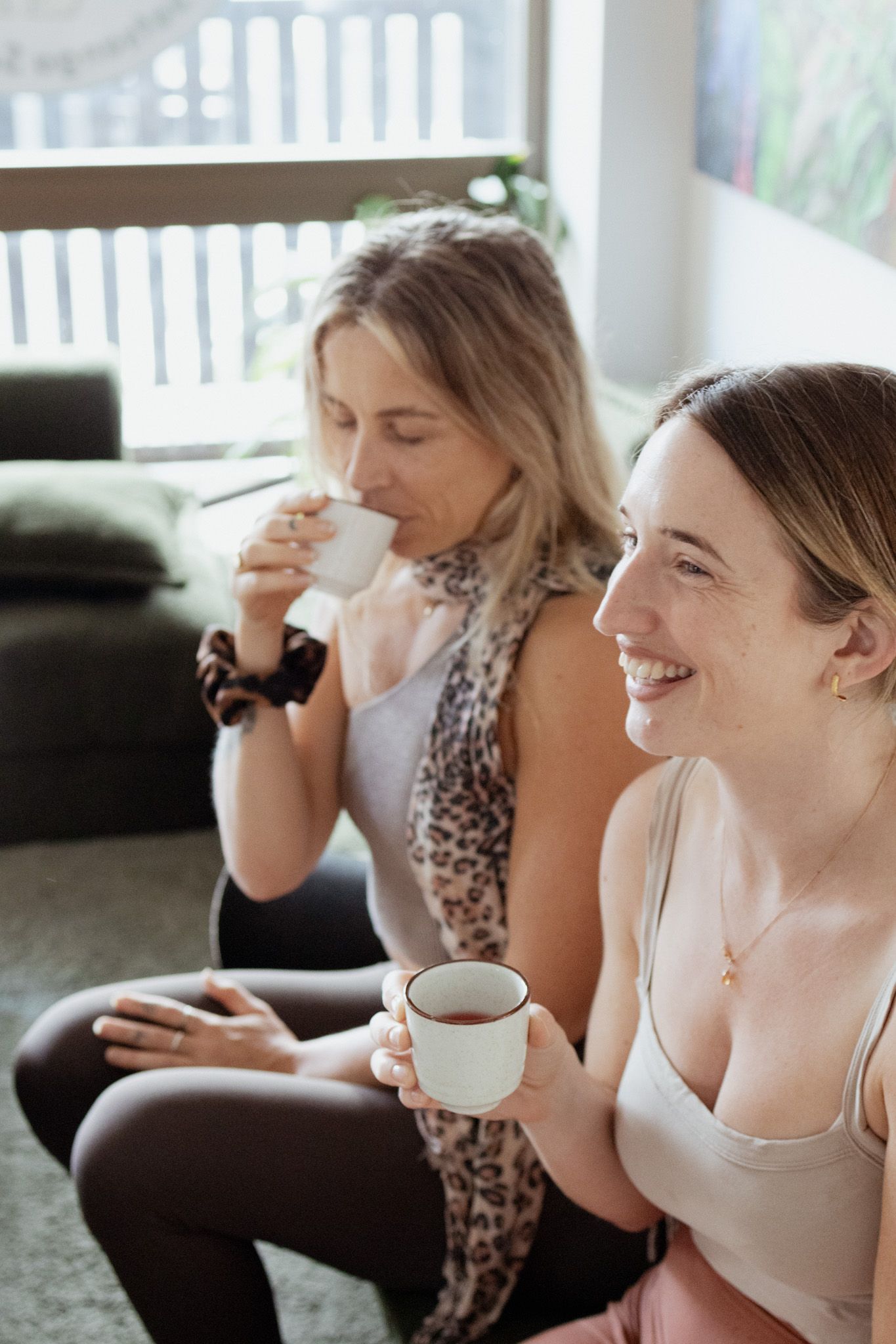 Two women sit together on a living room floor, smiling and sipping tea from small cups in a bright, cozy home setting.