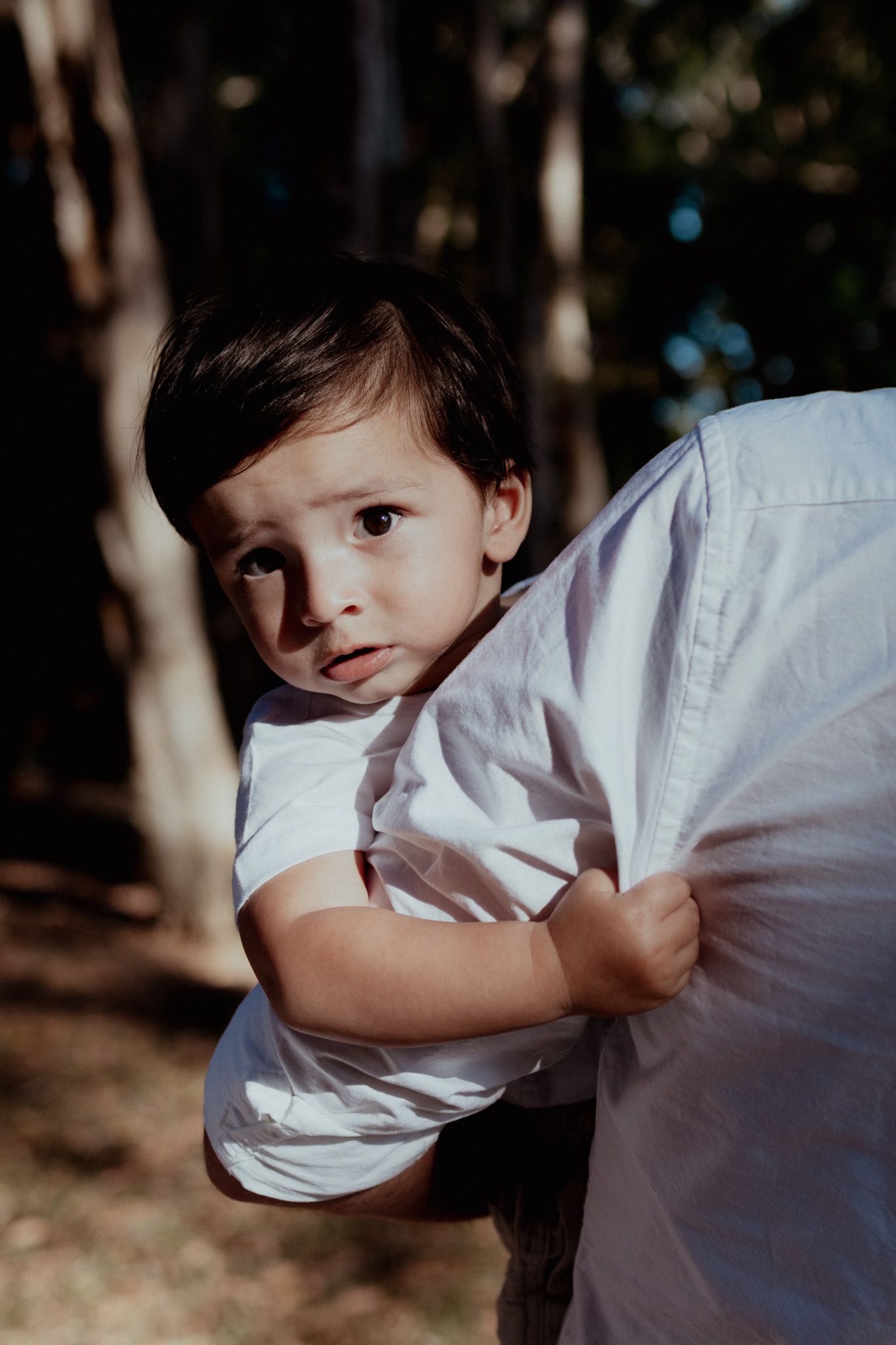 Young child in white shirt peeks around while being held outdoors.