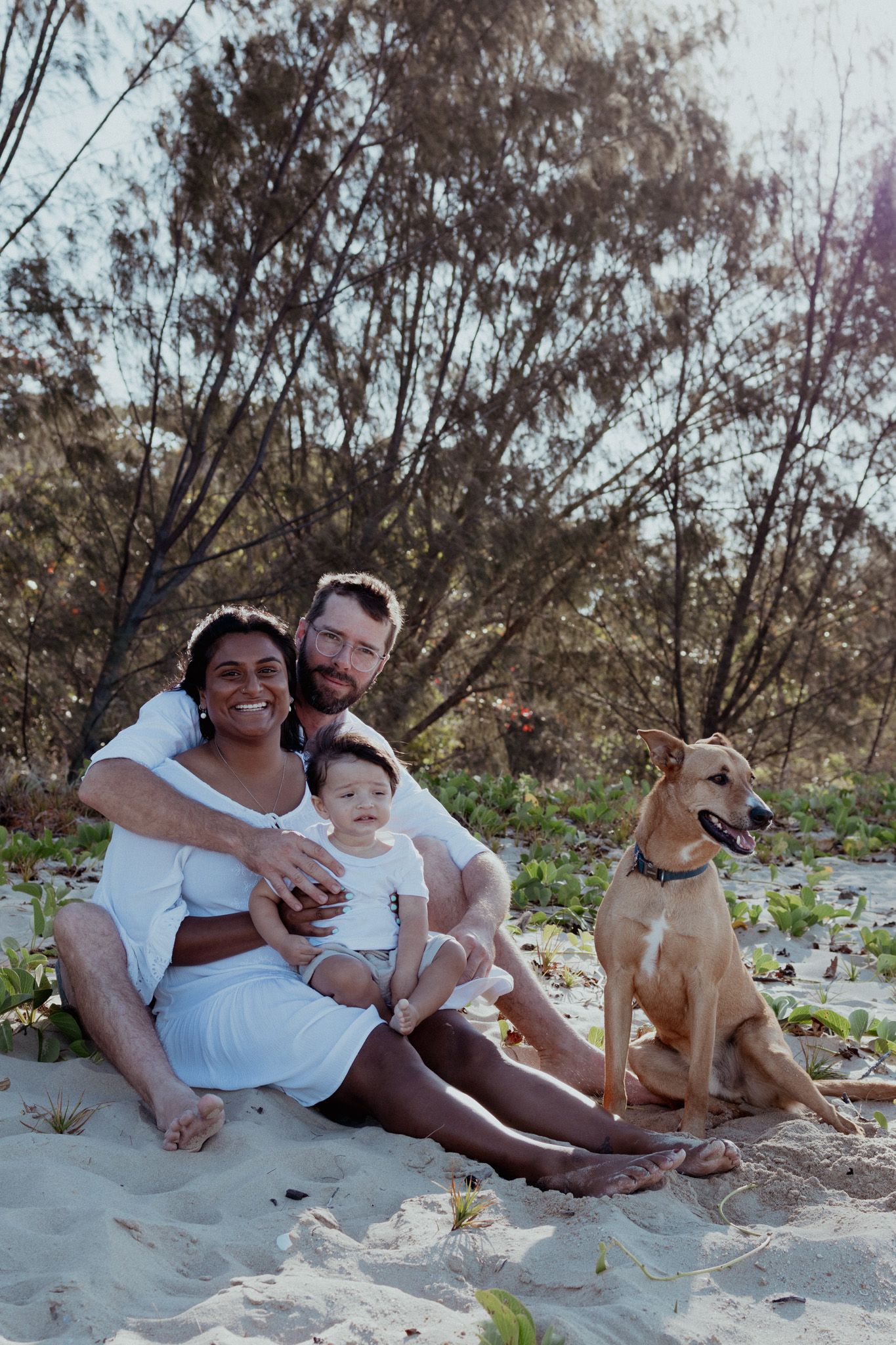 A casual beach photoshoot featuring a family relaxing on sand with their friendly dog.