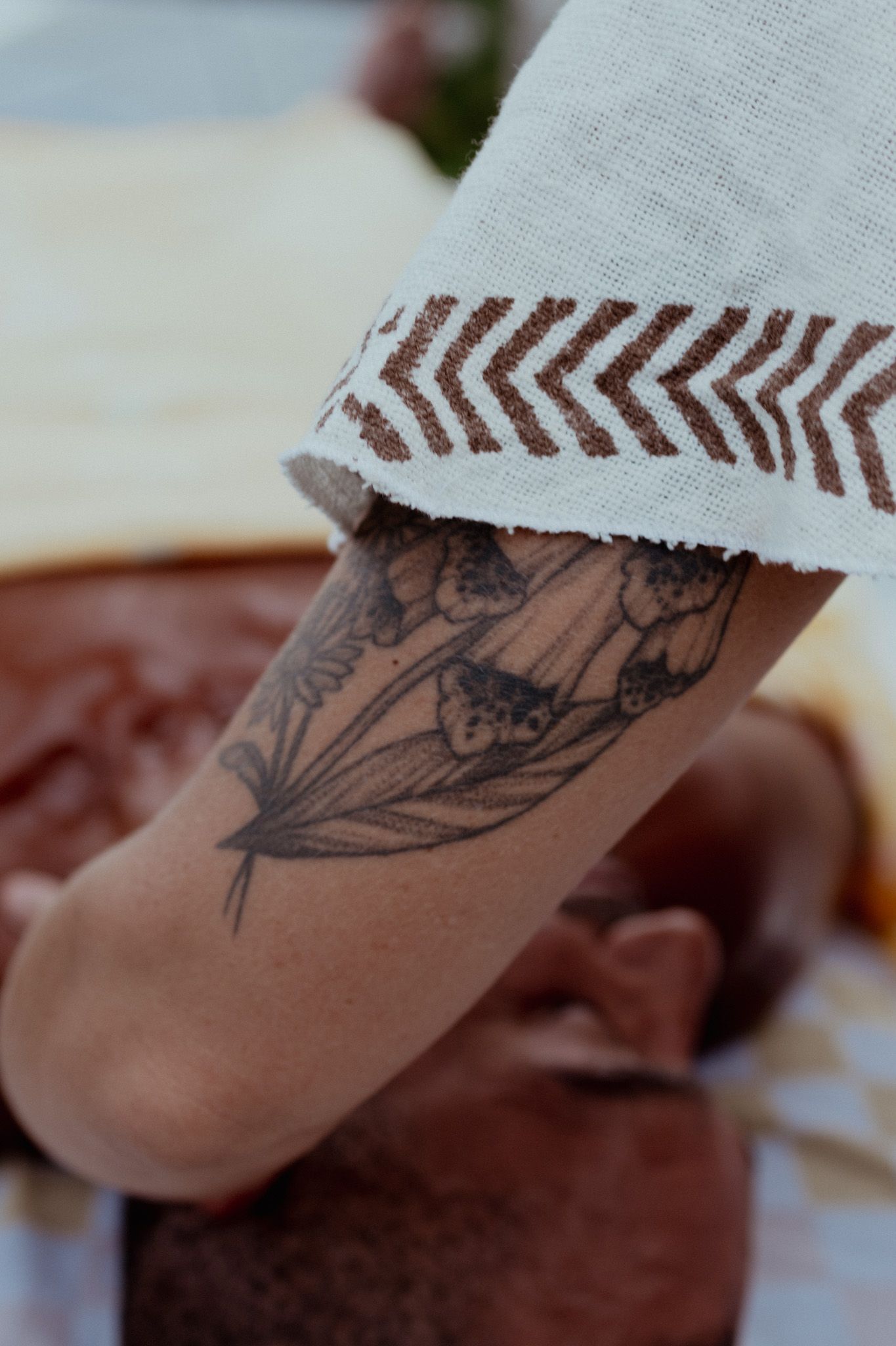 Close-up of a botanical floral tattoo on an upper arm, with a white sleeve showing a red chevron pattern and a person lying nearby.