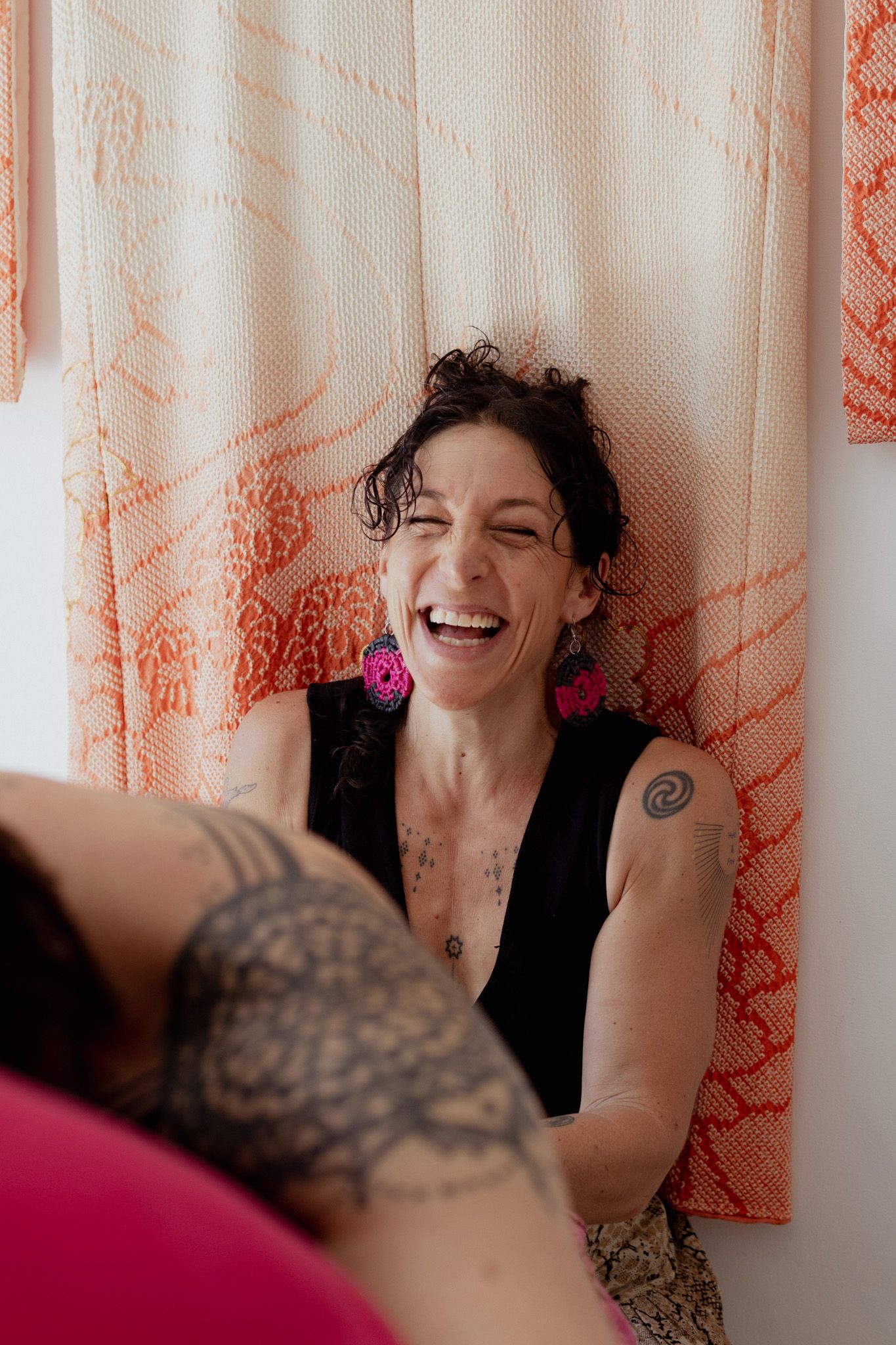 A laughing woman with dark curly hair, pink earrings and tattoos sits in front of orange patterned hanging textiles beside a person in lace.