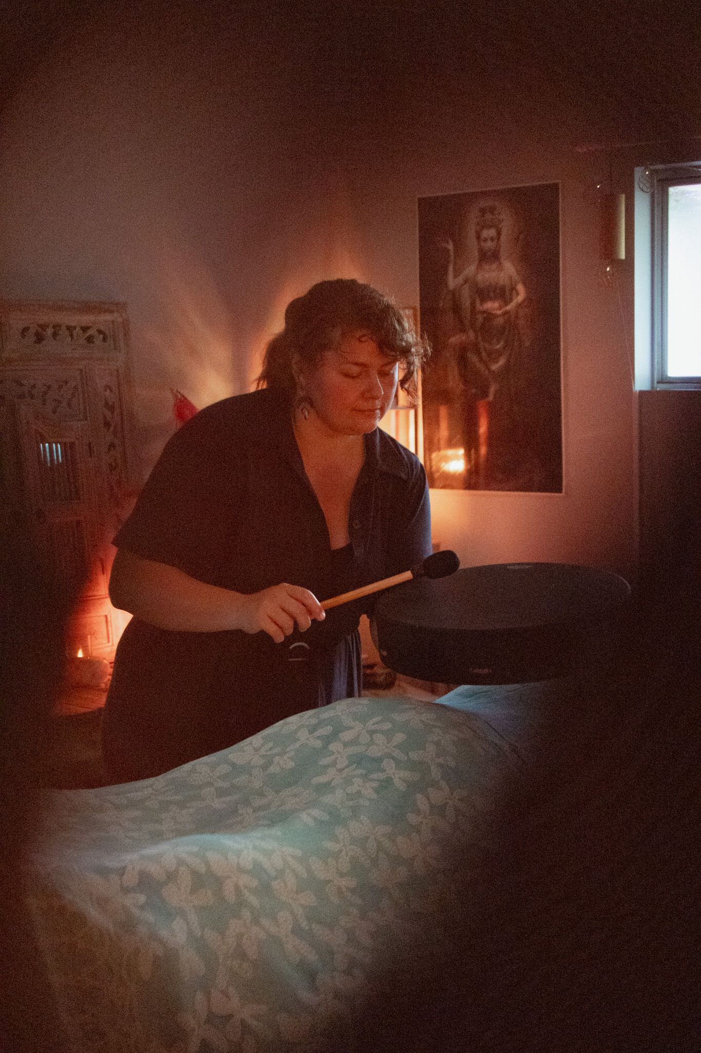 Woman holding a mallet over a large drum beside a massage table in a candlelit room with a spiritual painting on the wall.