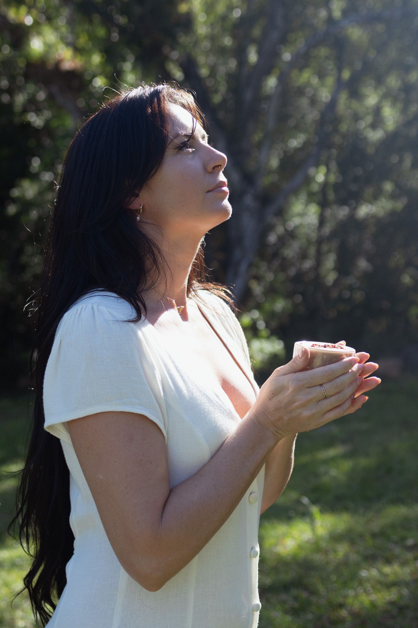 Woman with long dark hair in a white blouse gazes upward, holding a small cup in cupped hands, bathed in sunlight among green trees.