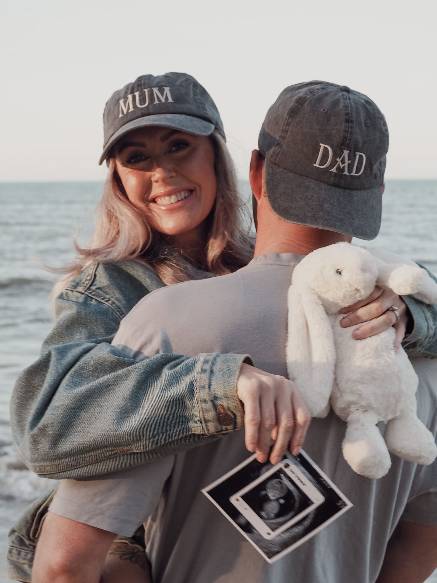 Couple wearing MUM and DAD caps holding ultrasound photo and stuffed bunny at beach.