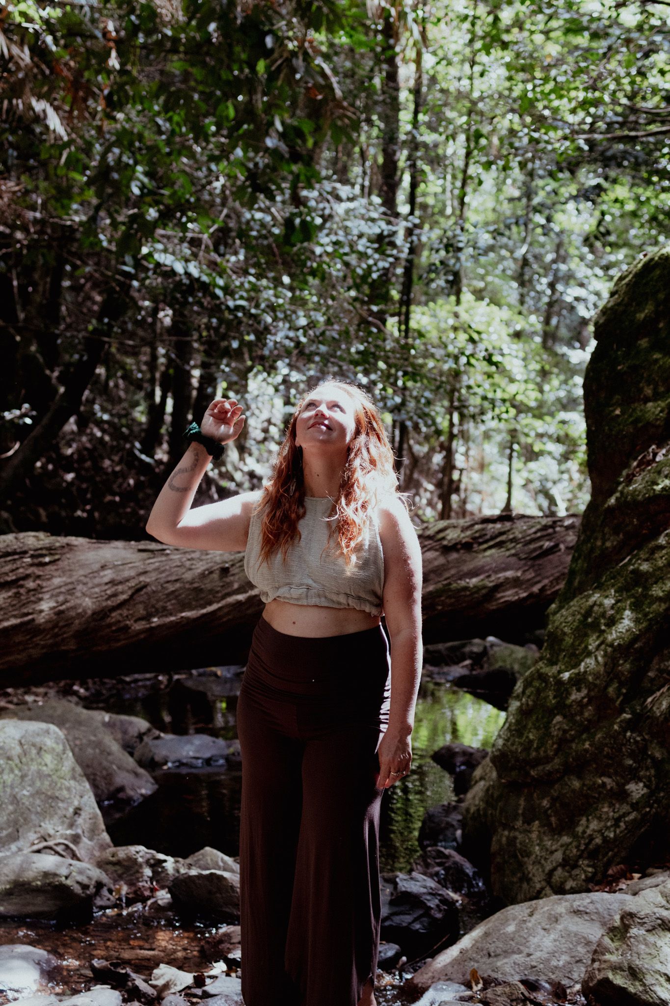 Figure in flowing dress poses among mossy rocks and trees in a forest setting.