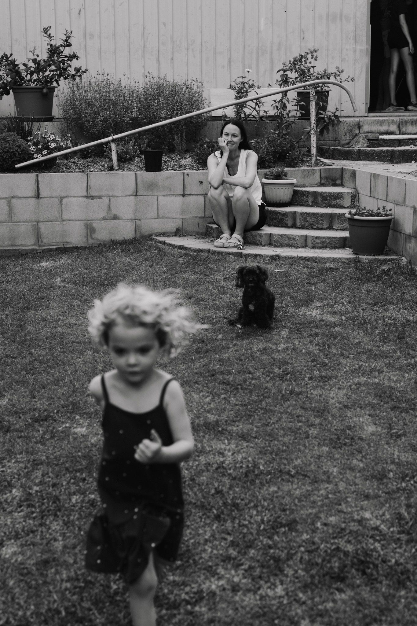Black and white photo of a backyard scene with a small child running and a black dog sitting on the lawn.