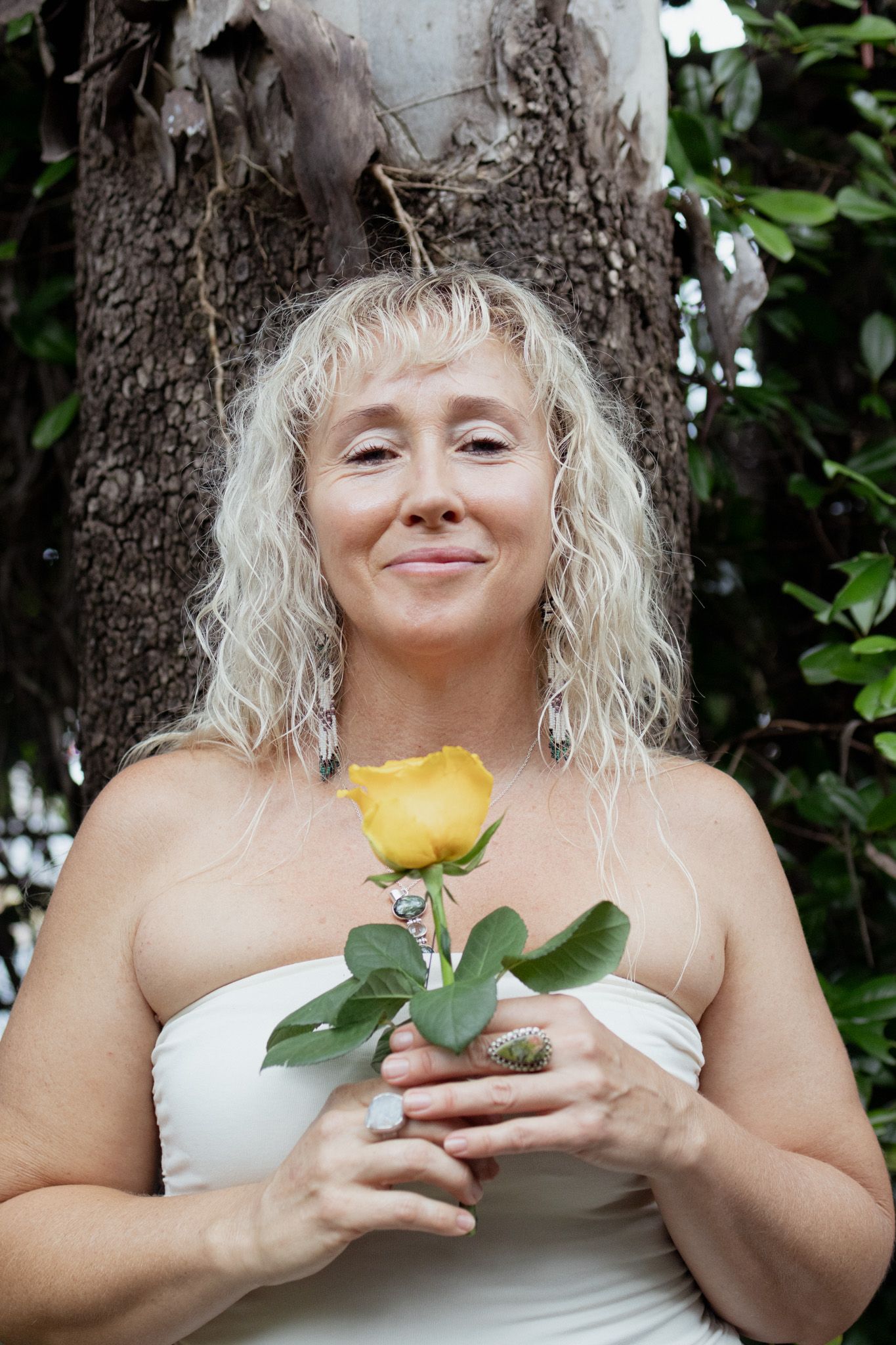 Woman with long curly blonde hair and a serene smile holds a yellow rose beside a tree trunk, wearing a white strapless top and rings.
