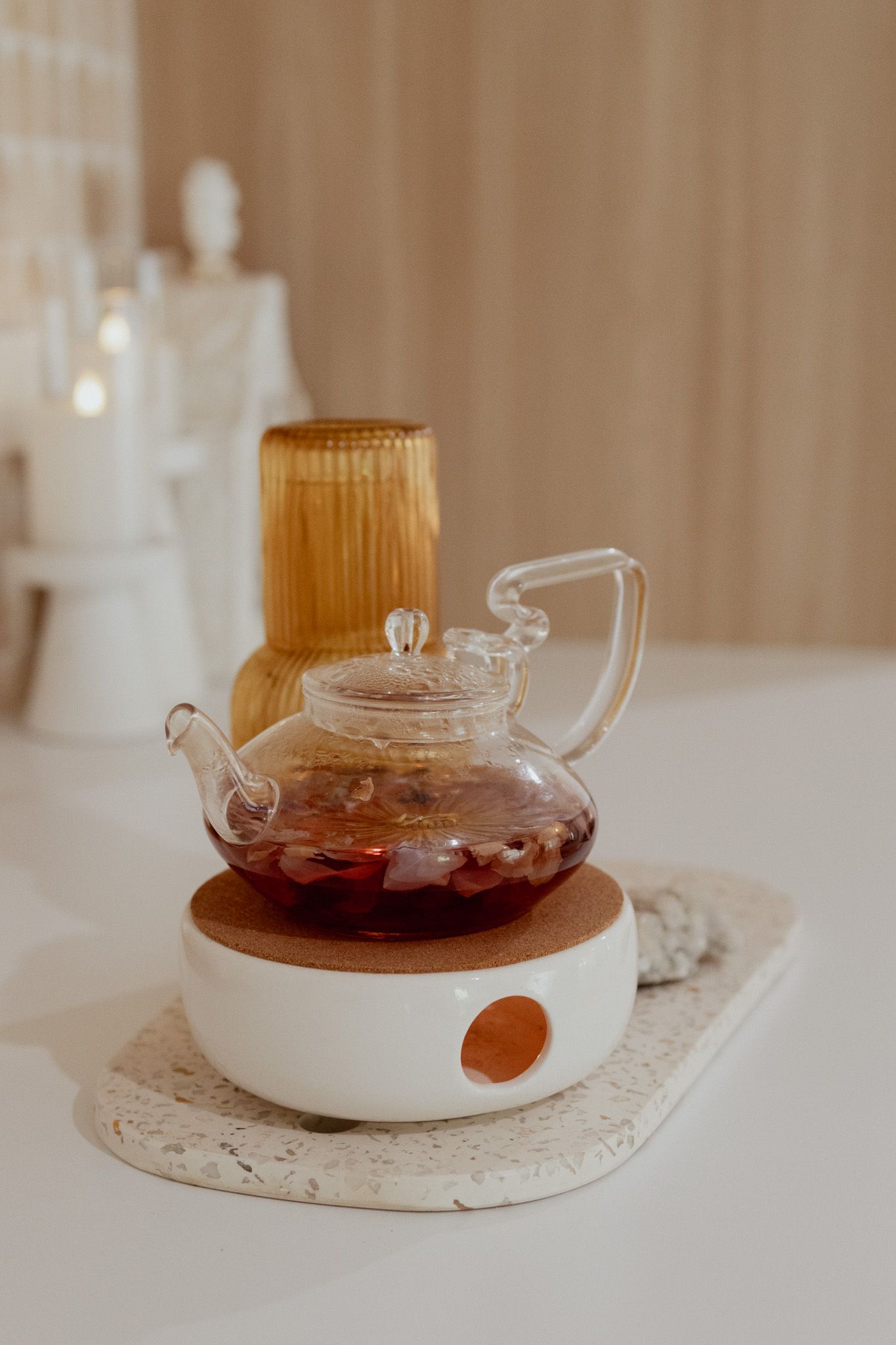Glass teapot filled with red fruit tea sits on a white ceramic warmer with cork top, beside an amber glass tumbler and candle.