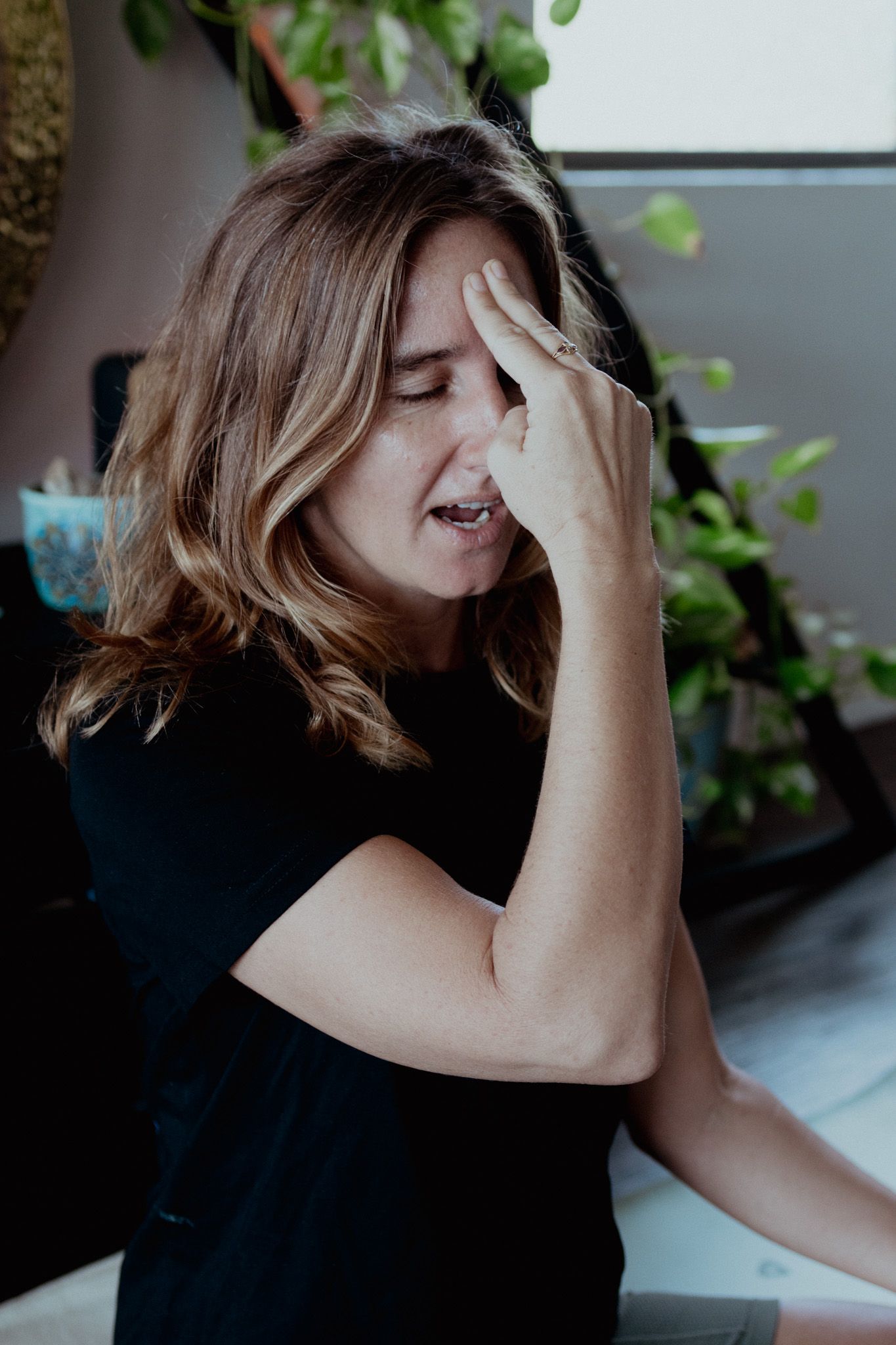 Woman in black top laughing with hand on face in indoor setting with plants.