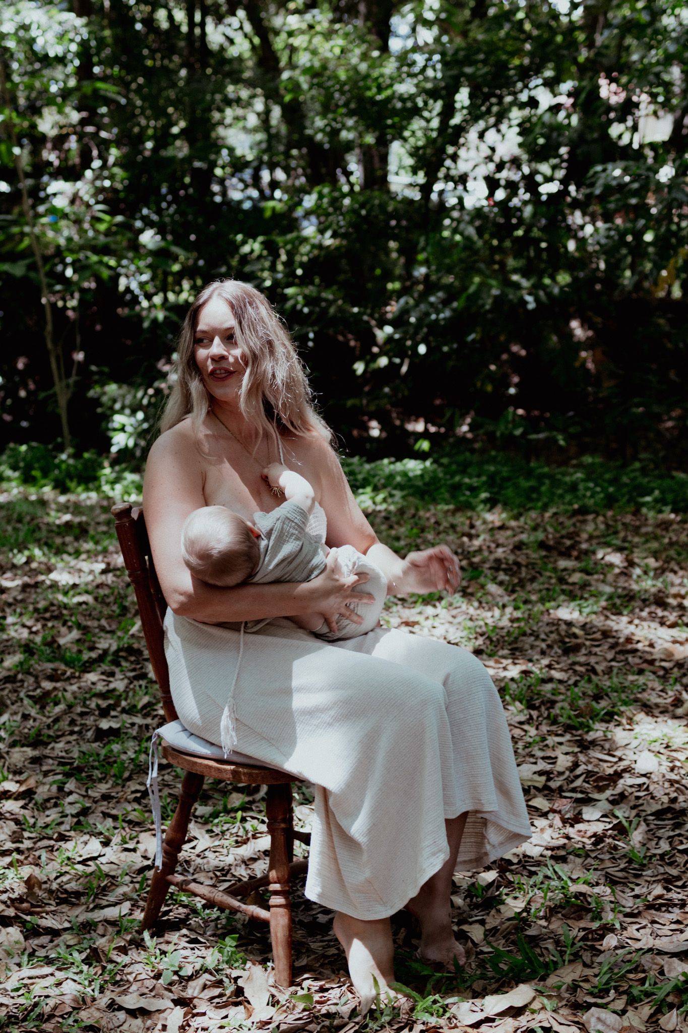 A serene outdoor portrait of someone in a white dress nursing a baby while seated on a wooden chair in a forest setting.