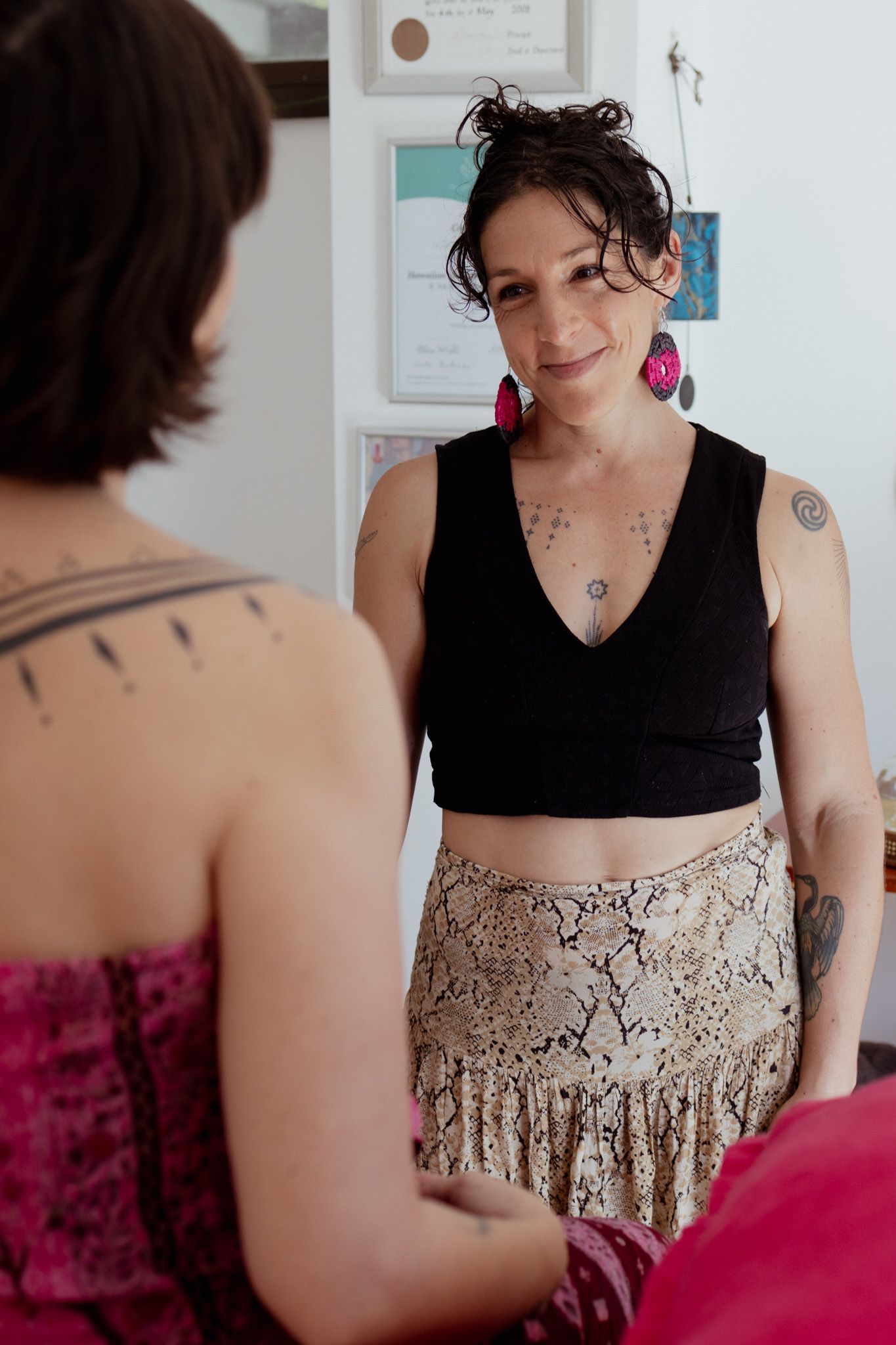 Smiling woman with tattoos, pink earrings, black crop top and snakeskin-print skirt facing a woman with short dark hair in a pink top.